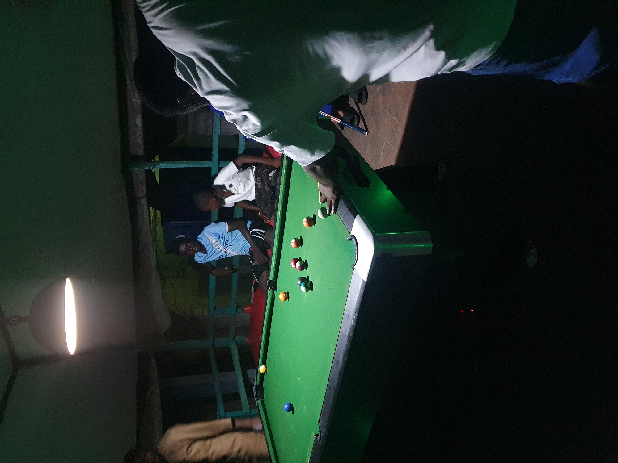 A group of people are gathered around a pool table in what appears to be an outdoor or semi-outdoor establishment in Lokoja, Nigeria. The lighting is dim, with a prominent overhead lamp illuminating the pool table. One individual, seen from behind and wearing a light-colored shirt and blue shorts, is actively playing pool, with their hand on the cue stick. Other individuals, including two young boys and an adult man, are visible in the background, observing the game or engaged in conversation. The pool table is green with visible balls scattered across its surface. The environment suggests a casual recreational setting, possibly a local bar or entertainment spot.