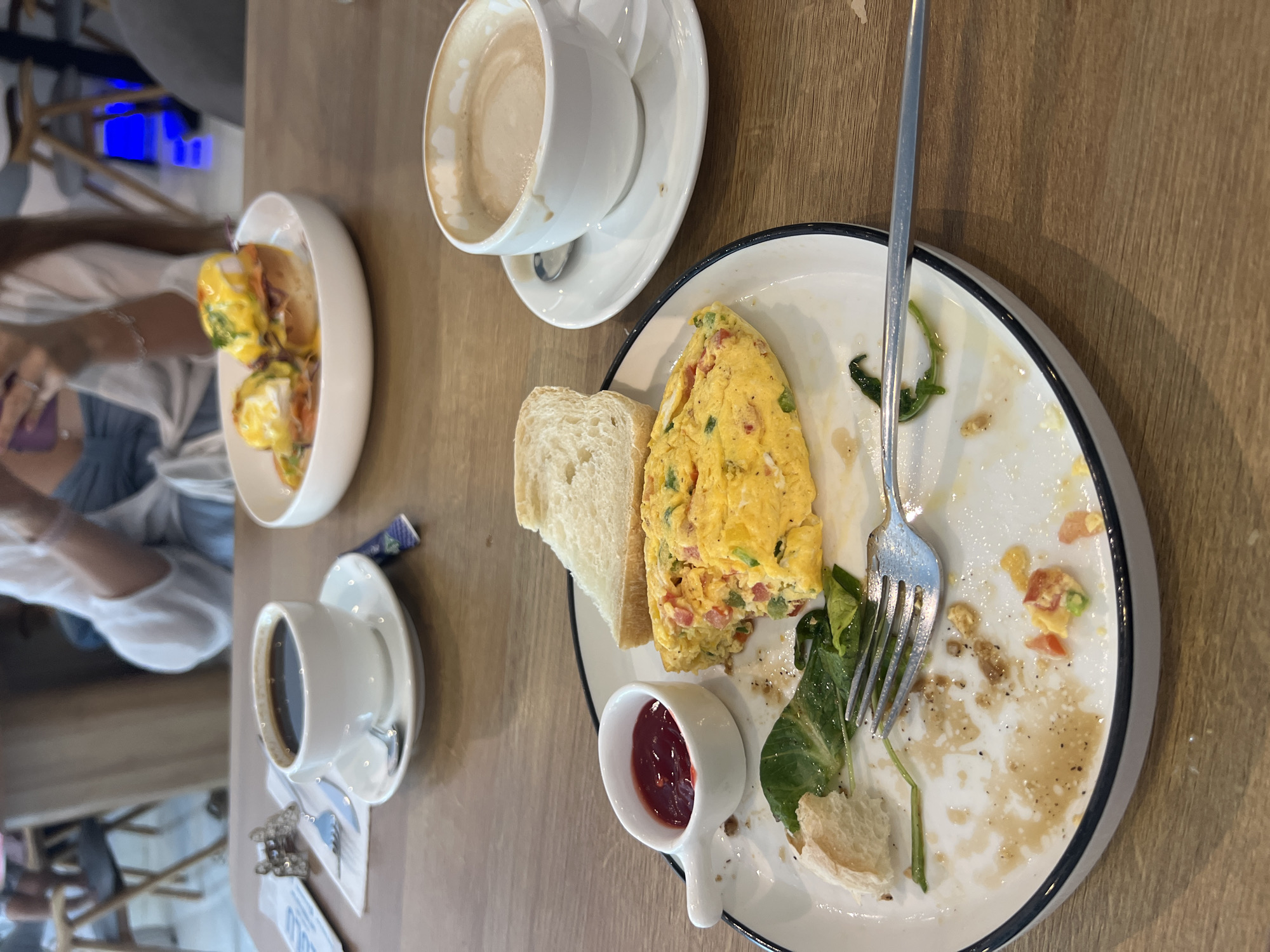The image shows a partially eaten breakfast on a light brown wooden table. The food consists of a half-eaten omelet, two slices of white bread, and a small container of red sauce on a white plate with a dark blue rim. A silver fork rests on the plate. To the left of the plate is a white bowl containing two eggs benedict. A white cup of coffee with a saucer is positioned above the plate, and another white cup of black coffee with a saucer is below it. A portion of a person's arm and torso are visible on the left side of the frame, along with chairs. The location is likely a cafe or restaurant in Bắc Nha Trang, Vietnam, suggested by the style of food and the overall ambiance.