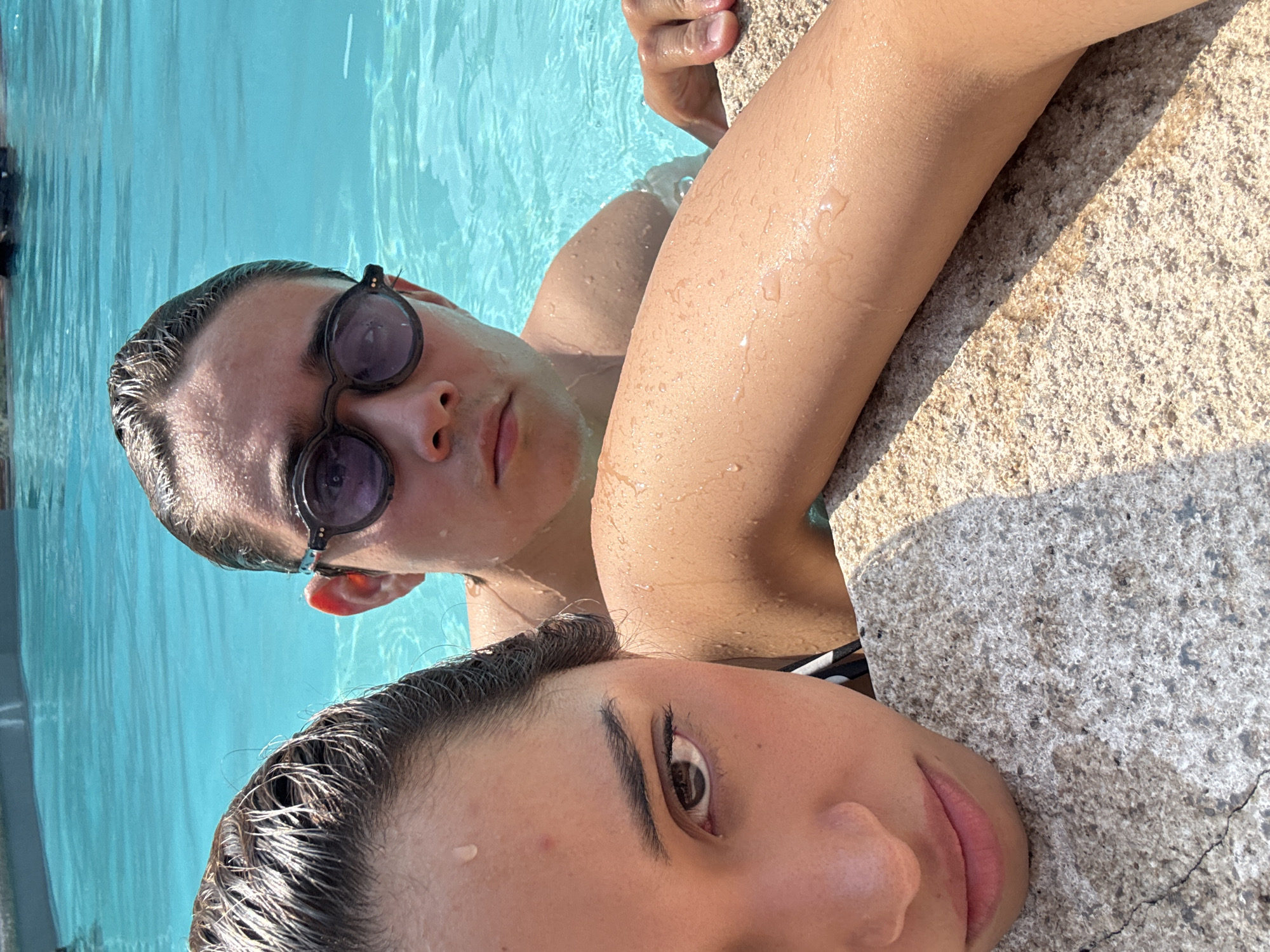 The image shows two people in a swimming pool. The location is Cocoyoc, Mexico. One person, wearing sunglasses, is partially submerged, looking at the camera. The other person is closer to the foreground, with their head resting on the pool's edge. Water droplets are visible on their faces and hair. The pool water is a clear blue. The pool edge is made of rough, light-colored stone.