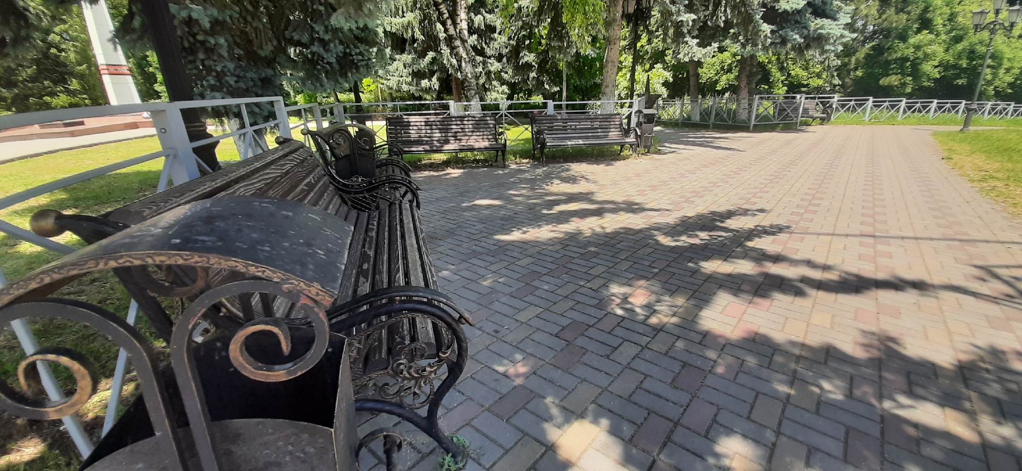 This is a photograph of a park in Yessentukskaya, Russia, on a sunny day. In the foreground, a wrought-iron park bench with a wooden seat is visible, angled towards the right of the frame. The bench has decorative scrollwork on its arms and base. Beyond the bench, a paved walkway, made of multi-colored bricks, extends into the distance. Trees with lush green foliage line the walkway on the left and right. In the background, another park bench and a trash receptacle are situated near a white metal fence. Further back, a tall, white monument with a red inscription stands partially obscured by trees. Sunlight casts dappled shadows of the trees onto the walkway, indicating a bright and clear day.