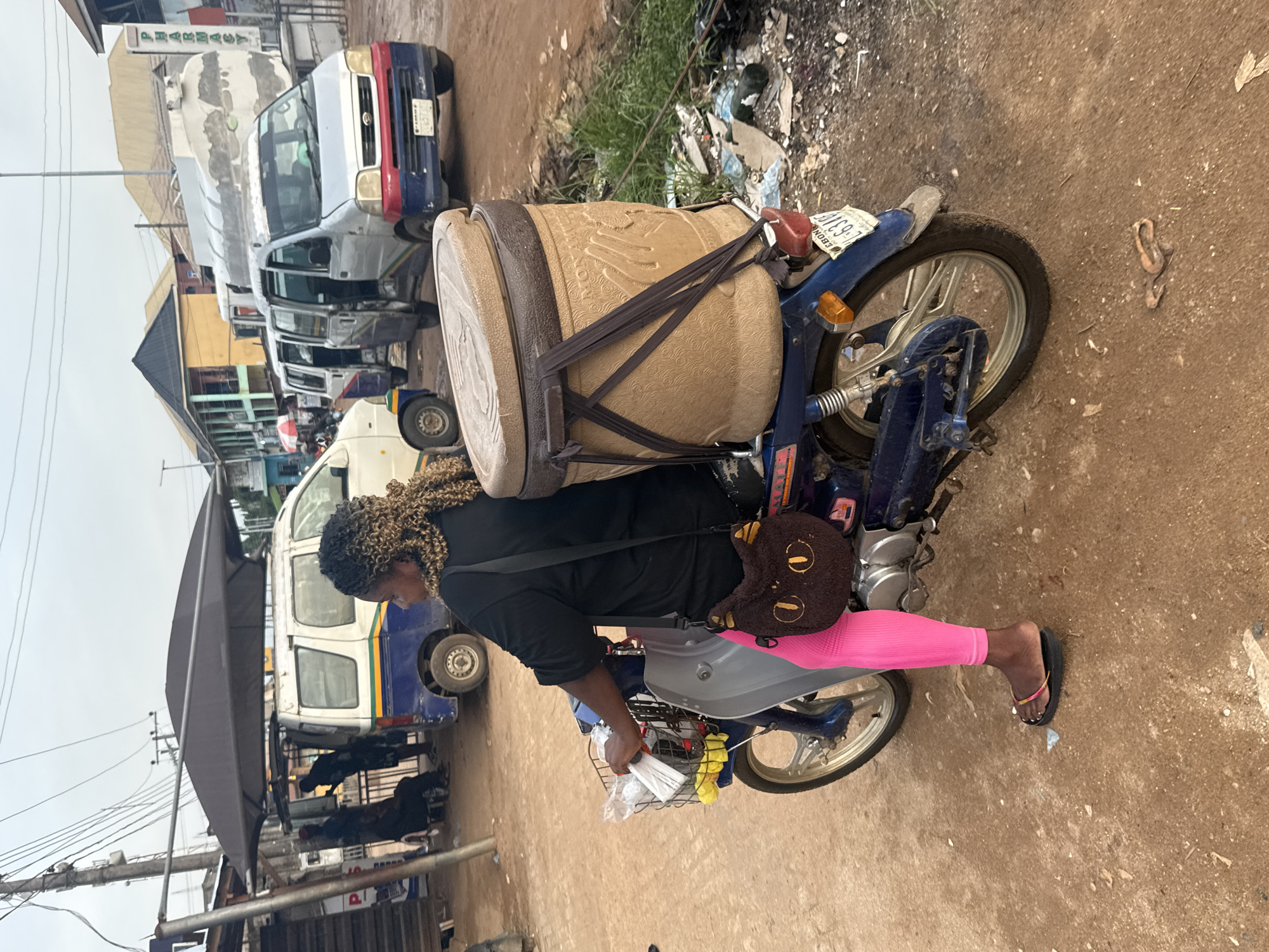 The image captures a bustling street scene in Ihiagwa, Nigeria, focusing on a person riding a motorcycle. The central subject is a person, seen from the side, with curly brown hair, wearing a black short-sleeved top, bright pink leggings, and dark flip-flops. They are seated on a blue motorcycle that appears to be stationary or moving slowly.
The motorcycle is heavily laden with cargo. A large, light brown, drum-shaped container with intricate embossed patterns is securely fastened to the rear seat with multiple dark straps. A small, round, brown purse with a smiling face design hangs over the rider's shoulder. In front of the rider, a small metal basket is attached, holding several white, individually wrapped items, possibly snacks or other goods.
The background reveals a typical street environment. To the left, several vehicles, including vans and cars, are parked or driving. One prominent van has a distinctive red and blue front. Further back, buildings with awnings suggest shops or market stalls, and a sign with the word "PHARMACY" is clearly visible. Utility poles and overhead wires crisscross the sky. The ground is an unpaved, dusty, reddish-brown surface with some scattered debris and sparse vegetation. The sky is overcast, indicating daytime but not bright sunshine. A visible license plate on the motorcycle's rear fender reads "EBONYI," likely referring to Ebonyi State in Nigeria.