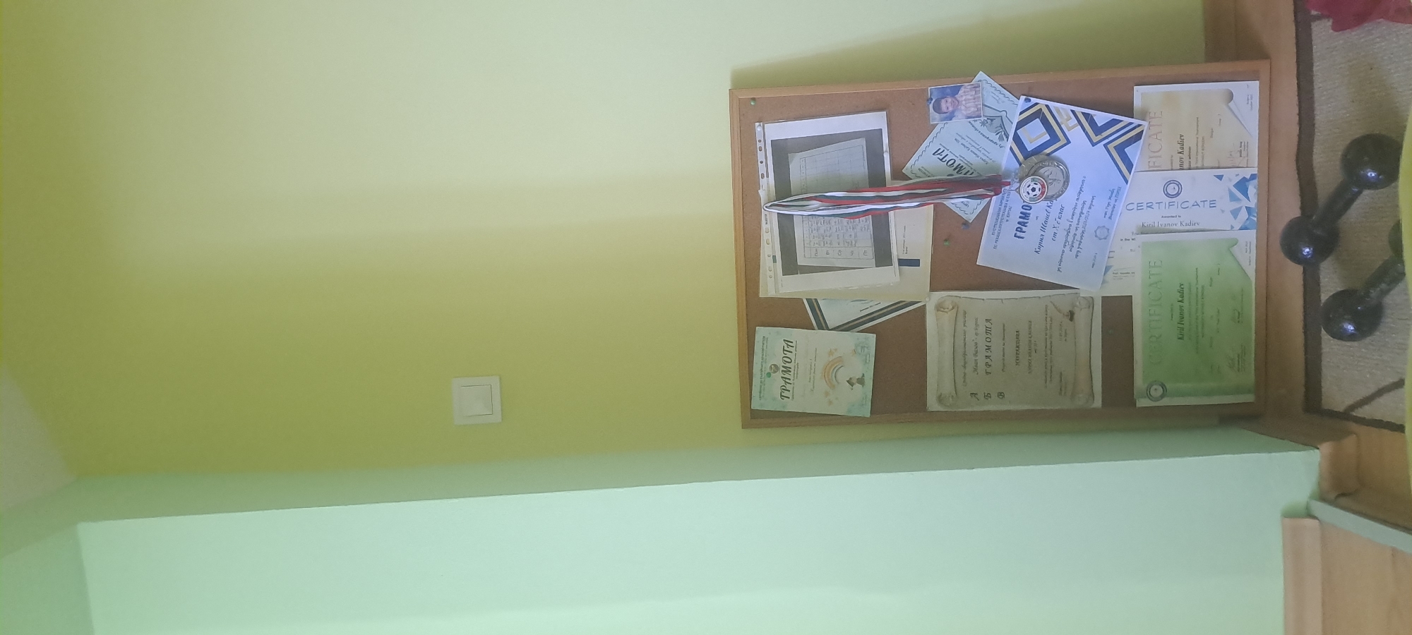 The image shows a bulletin board with several documents and a medal. The bulletin board is mounted on a wall painted yellow. To the left of the bulletin board is a wall painted a lighter green. On the wall, above the bulletin board, is a white light switch. The bulletin board contains multiple certificates and a medal with a ribbon. One certificate is green and has text that includes "CERTIFICATE" and "Kiril Ivanov Kadiev". Another certificate is white with a crest and text in Bulgarian that appears to be "ГРАМОТА". A medal hangs from the bulletin board, attached to a red, white, and blue ribbon. A small framed photograph of a young boy is also pinned to the board. Below the bulletin board, two dumbbells are visible on the floor. The scene appears to be an indoor space, possibly a home or a sports facility. The context of Burgas, Bulgaria is not directly visible in the image itself, but assuming this is a personal bulletin board in a residence within Burgas, it likely represents achievements or recognitions related to the individual named on the certificates.