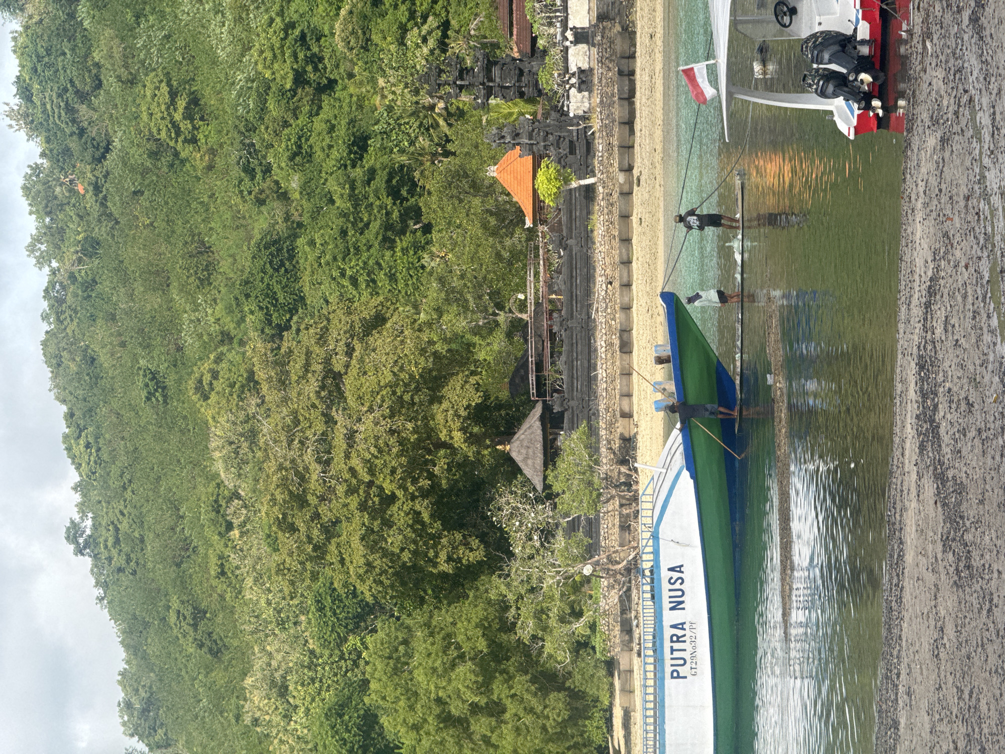 The image shows a coastal scene in Nusa Penida, Indonesia. Lush green vegetation covers the hillside. At the shoreline, there are structures, including a building with an orange tiled roof, indicating a residential or public area. A boat named "PUTRA NUSA" is partially visible in the water, along with other boats further out. Two people are standing in the shallow water near the boats. The shoreline consists of a sandy area and a concrete or stone barrier. The water is calm and reflects the sky and surroundings. The weather appears overcast.