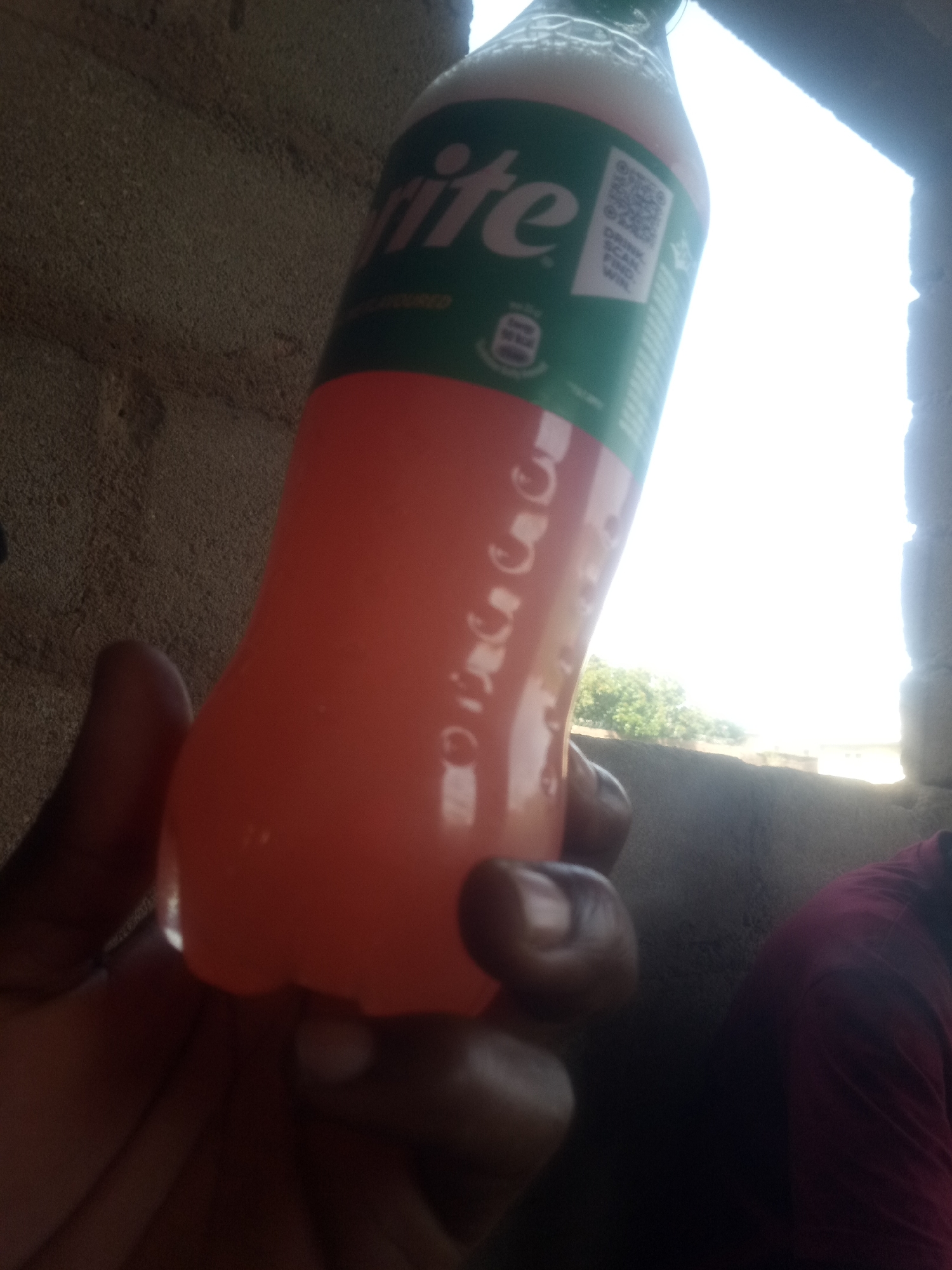 An over-the-shoulder shot shows a person holding a plastic bottle of Sprite with a pinkish-orange liquid inside. The bottle's label, predominantly green, is partially visible, featuring the Sprite logo and a QR code. The background reveals a textured, light brown wall and a bright, blown-out window that looks out onto a sunny outdoor scene with green foliage. A sliver of a person wearing a maroon shirt is visible in the lower right corner. The lighting suggests it is daytime. The image is slightly out of focus, particularly the background.