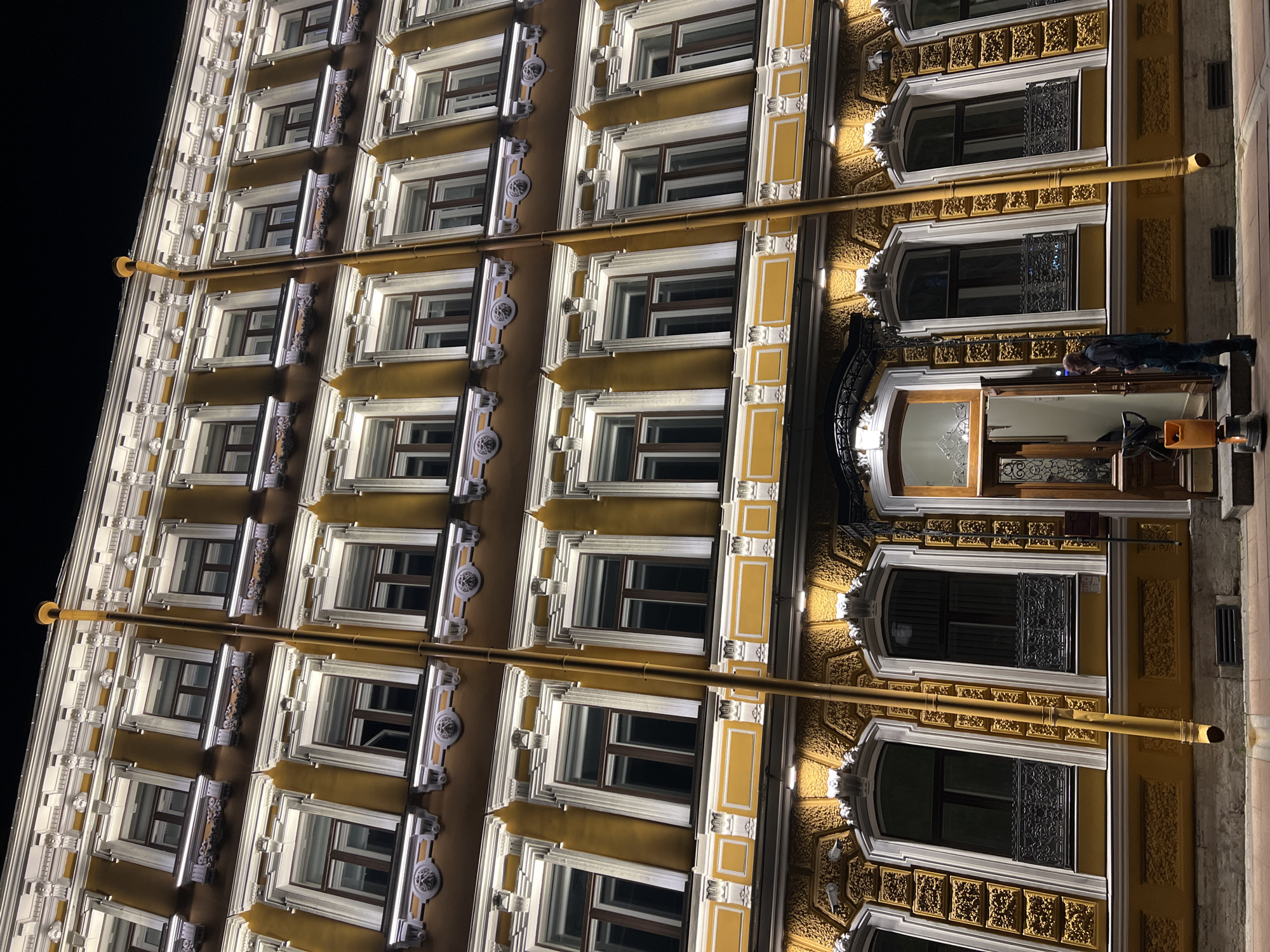 The image depicts an upward, angled view of a multi-story building, likely in Saint Petersburg, Russia, given the architectural style. The building has numerous windows, each framed with ornate decorative molding in white and gold. A yellow drainage pipe runs vertically down the facade. A person is seen standing in the doorway, engaged in an interaction with a door or object inside the building. The surrounding environment appears to be dark, suggesting the photo was taken at night.