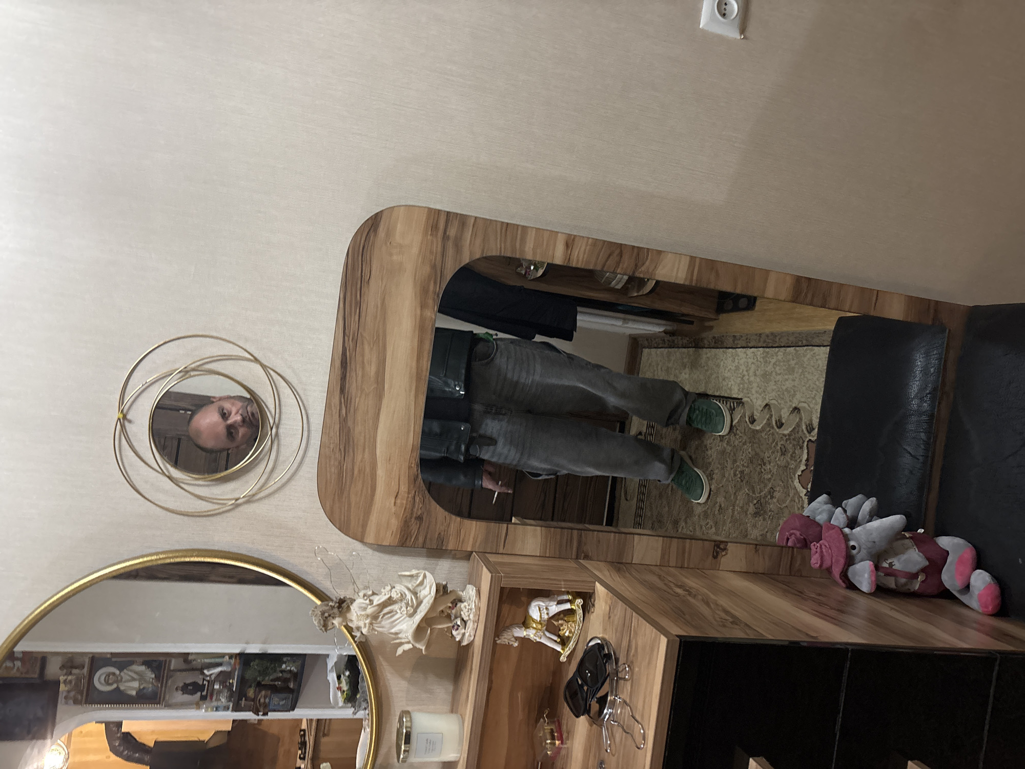 A person's reflection is visible in a mirror on a wooden cabinet. A circular mirror is on the wall, with the head of a man reflected in it. A larger, gold-framed circular mirror sits below. On the cabinet are decorative objects, including an angel figurine and a rocking horse. Next to the cabinet is a black leather chair with a stuffed animal on top. The scene is likely in Tbilisi, Georgia, based on the context of the location.