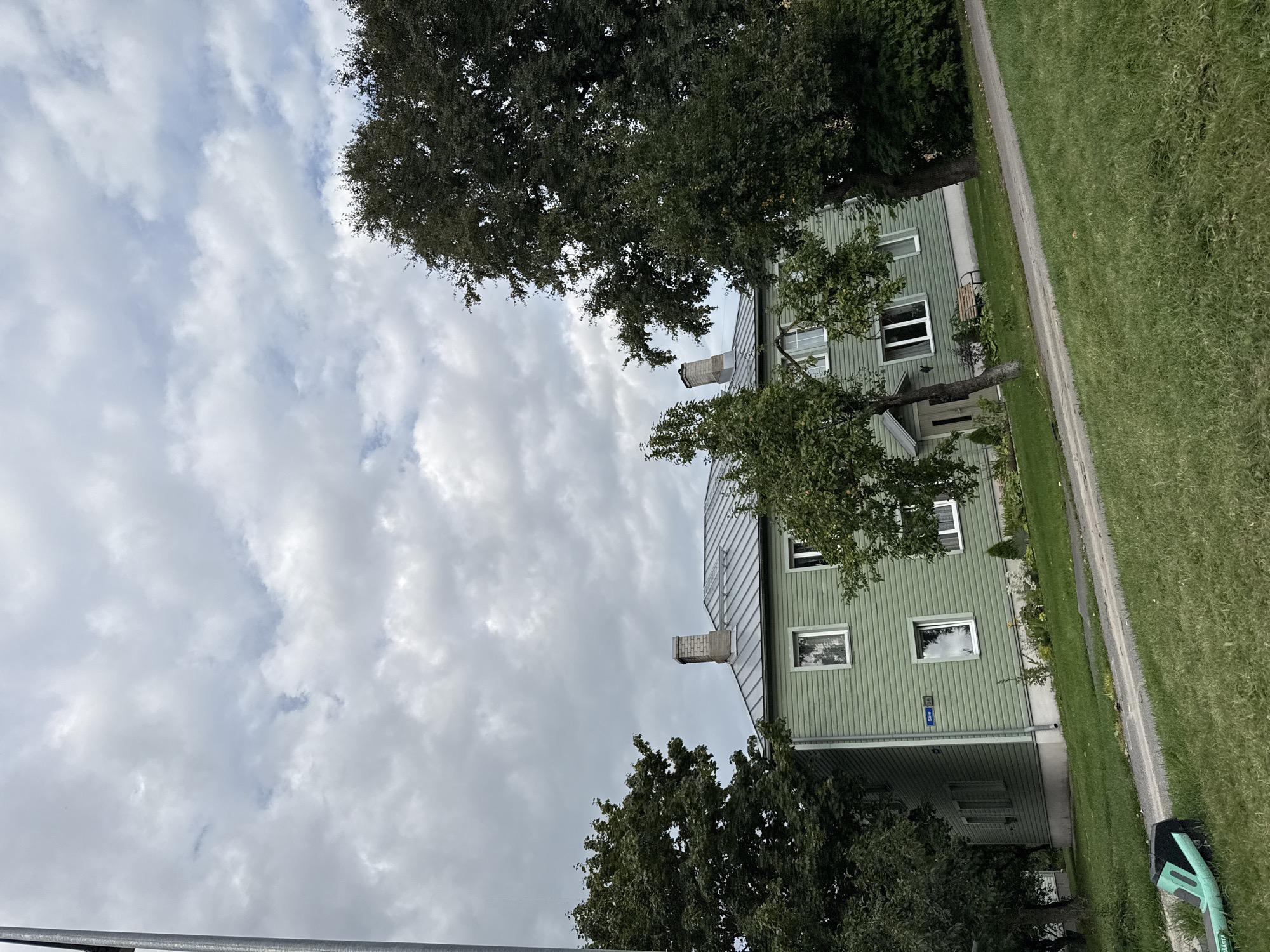 This is a photograph of a green, two-story building in Tallinn, Estonia. The building has a metal roof, multiple windows, and two chimneys. The house appears to be in a residential area with trees and grass surrounding it. A street sign that reads "Sõle 98" is visible on the side of the building. The sky is overcast with clouds. In the foreground, there is a green and black object that appears to be a scooter or a similar recreational vehicle. There are no people visible in the image. The photograph is taken from a slightly elevated angle, looking up at the building. The building is likely an apartment building or a duplex given its size and the presence of multiple windows on each floor. The environment appears to be a typical suburban or urban residential setting in Estonia.