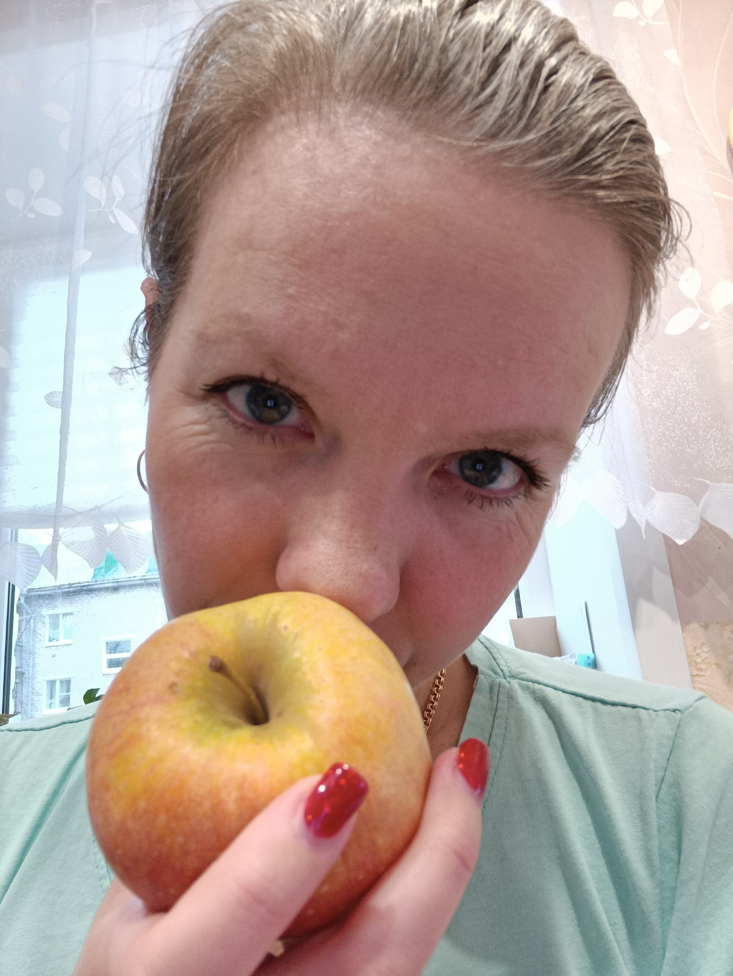 A close-up, eye-level portrait shows a woman holding a yellow and orange apple to her nose/mouth, looking directly at the camera. She has fair skin, blue-green eyes, and light-colored hair pulled back from her face, revealing a high forehead. Fine lines are visible around her eyes. She wears a light mint-green t-shirt and a thin gold chain around her neck. Her fingernails are painted a vibrant red.
The background primarily features a large window with white, translucent curtains adorned with a subtle leaf pattern. Beyond the window, a light-colored brick building with multiple windows is visible, suggesting an urban or suburban environment in Chaikovsky, Russia. The natural light filtering through the window indicates it is daytime, possibly bright but not directly sunny. Inside, to the right, hints of light-colored walls or furniture and possibly a cardboard box can be seen. The overall mood is neutral and focused. There is no visible text in the image.