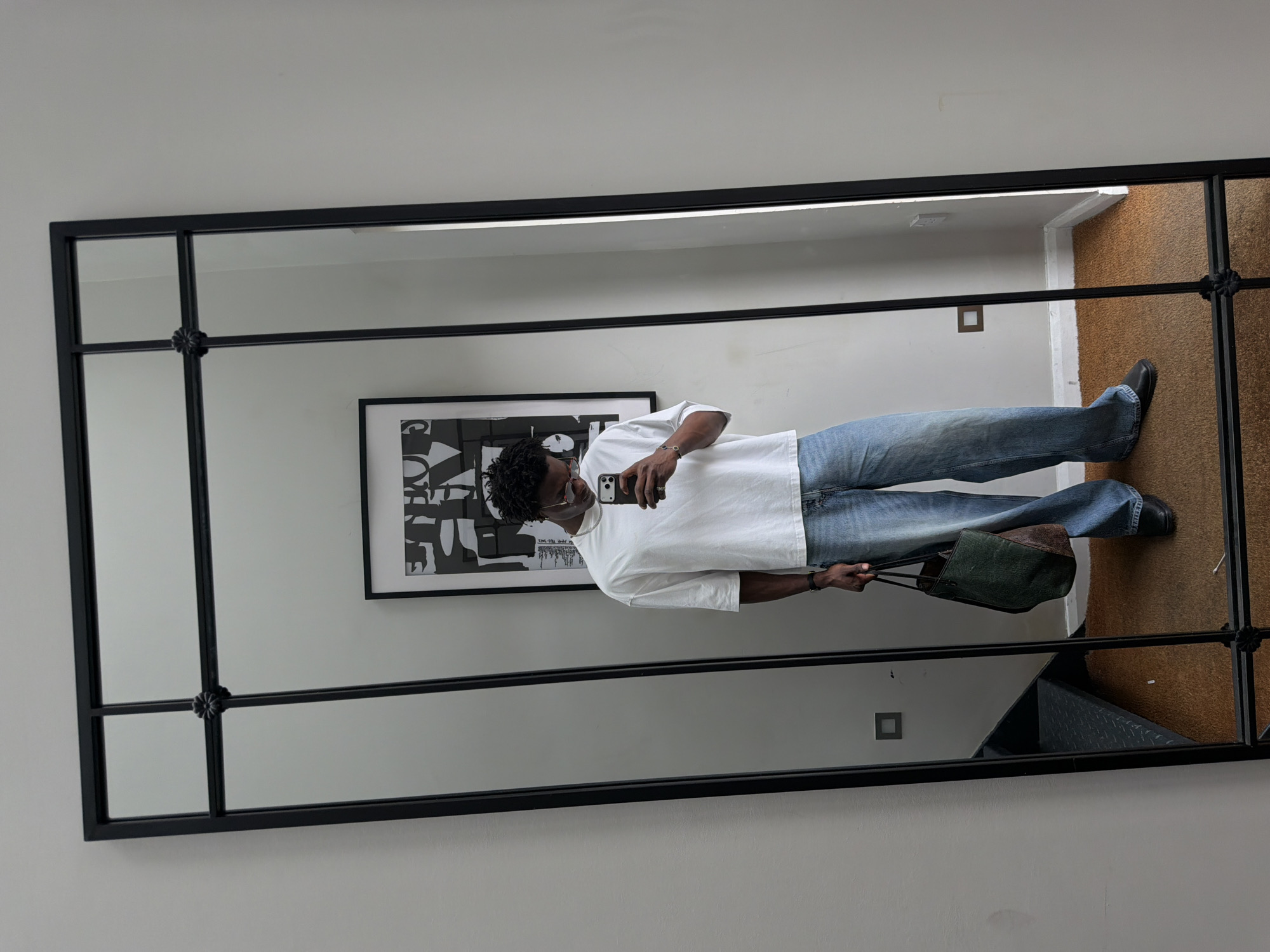 A man takes a mirror selfie in an interior space. He is wearing a white t-shirt, light blue jeans, black shoes, and headphones. He holds a phone in his right hand and a green and brown bag in his left hand. Behind him is a framed piece of artwork. The room has a light-colored wall and brown flooring, part of which appears to be a staircase. The location is likely an apartment in London, United Kingdom.