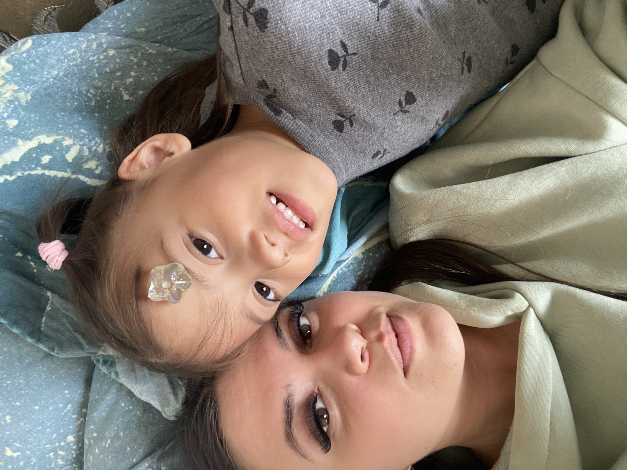 A close-up captures a woman and a young girl lying on a blue, patterned blanket. The girl, positioned at the top of the frame, has a decorative flower-shaped gem adhered to her forehead. A pink hair tie secures a small ponytail. She wears a gray sweater adorned with black floral designs. The woman, at the bottom of the frame, wears a light green hooded sweatshirt. The location in the city of Zhigalki, Russia is not discernable from the image.