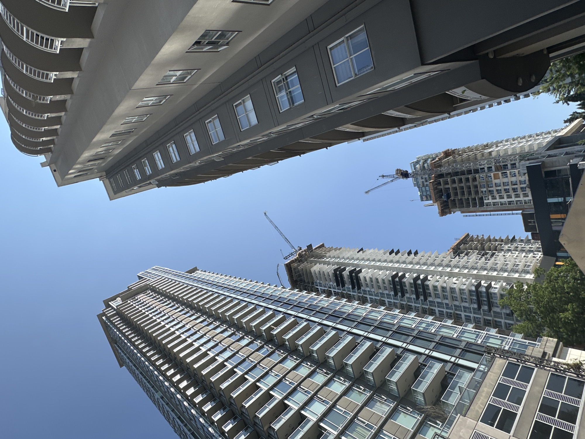 This image captures a low-angle view of several modern high-rise buildings against a clear blue sky. In the foreground, a dark gray, curved building with visible balconies dominates the right side of the frame. To the left, a sleek, glass-and-steel skyscraper with numerous balconies stretches upwards. In the middle ground, two tall buildings are under construction, indicated by exposed concrete frames and construction cranes. The building on the far left appears to be a completed residential tower with a glass facade and balconies. The scene is urban, likely a city center, and the presence of construction suggests ongoing development. The image was taken in Coquitlam, Canada.