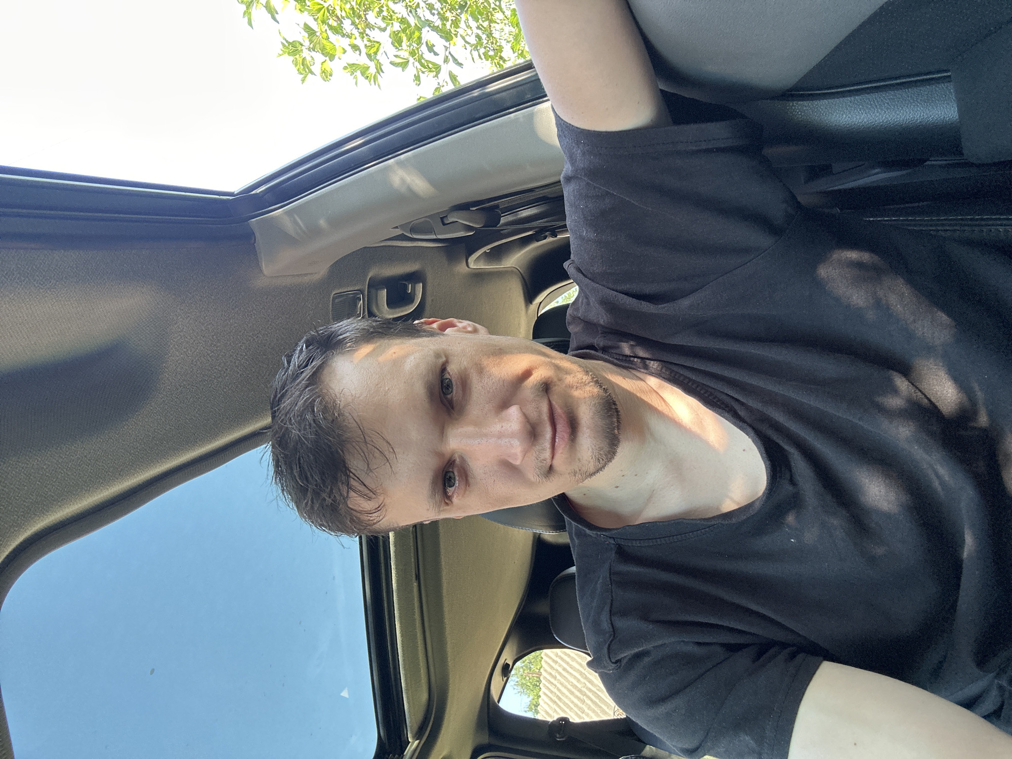 A man is taking a selfie in a car with a sunroof open. He is wearing a black t-shirt. The car interior is visible, including the seats and roof lining. Sunlight streams through the sunroof, casting shadows on the man's face and shirt. Outside the car, greenery and a fence are visible. The man appears to be looking directly at the camera.