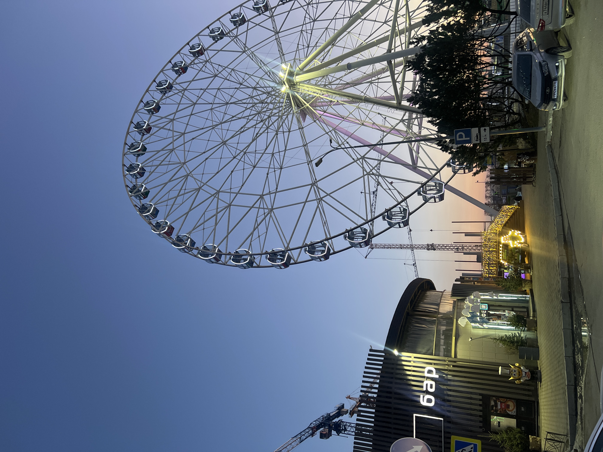 The image shows a large Ferris wheel in Kazan, Russia, likely the Kazan Riviera Ferris Wheel. The wheel is white with numerous enclosed passenger cars. The sky is a gradient of blue to orange/pink, suggesting twilight or dusk. A building with the word "бар" (bar) in Cyrillic script is in the foreground. Construction cranes are visible near the building and behind the Ferris wheel. Cars are parked along the street adjacent to the building. The image has an upward-tilted perspective.