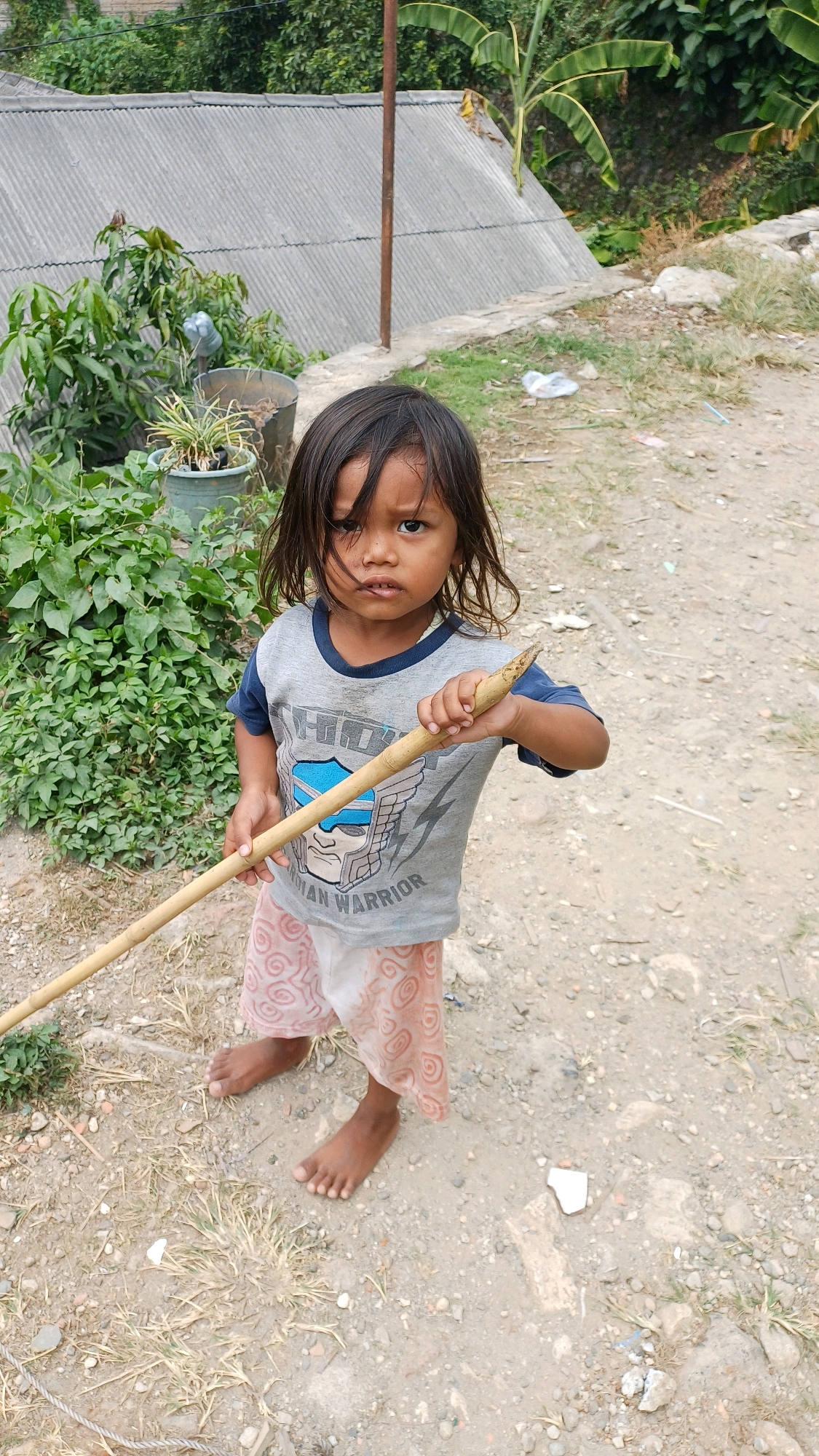 A young child stands outdoors, holding a wooden stick. The child is wearing a grey t-shirt with a blue collar and a pink skirt with a circular pattern. The child's feet are bare, and they are standing on a dirt path. In the background, there is a corrugated metal roof, green foliage, and some scattered debris. The setting appears to be a rural or semi-rural area, possibly in Citeureup, Indonesia, given the vegetation and structures.