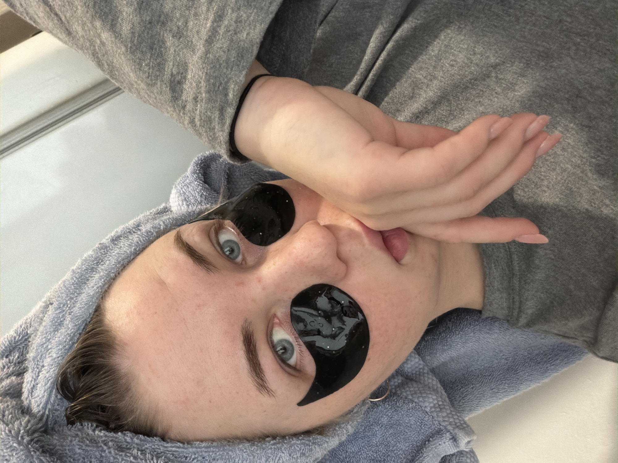 A person is lying on their back, with their head tilted to the right, wearing black under-eye patches. The person has light skin, blue eyes, and light brown hair that is partially covered by a blue towel. They are wearing a gray sweatshirt, and their right hand is near their mouth. The location of Chaikovsky, Russia, cannot be determined from the image.