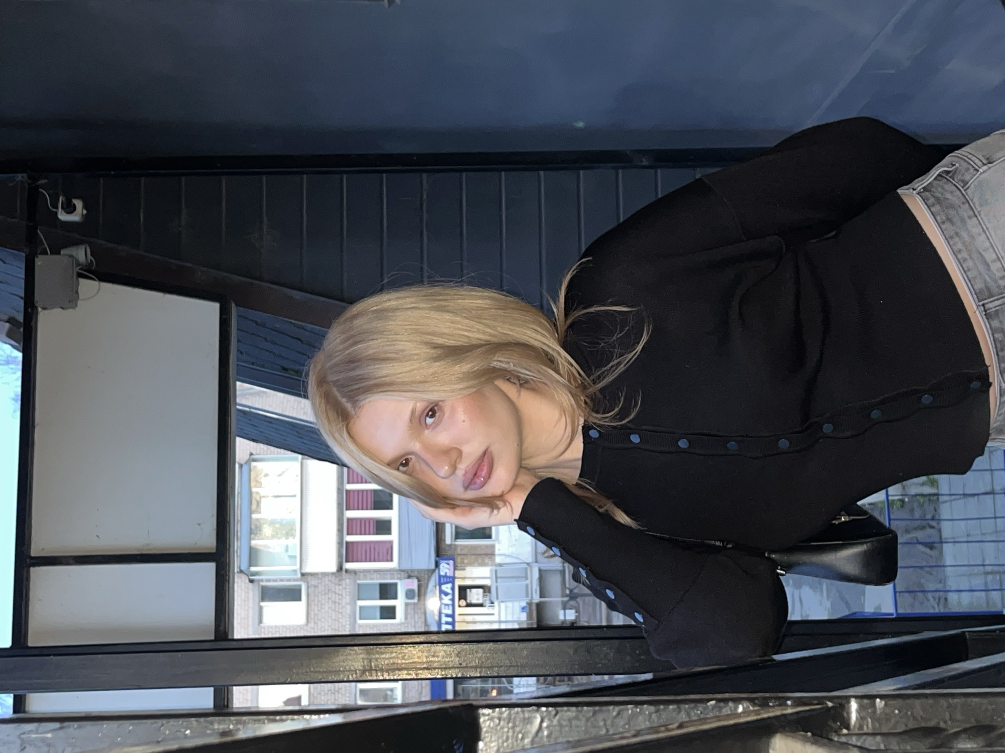 A head-and-shoulders portrait features a young woman with long blonde hair, dressed in a black cardigan with contrasting blue buttons and light wash denim jeans. She leans her head slightly, resting her chin on her left hand, and looks directly at the camera with a calm, neutral expression.
The setting appears to be an outdoor balcony or an elevated open area, with a dark metal railing framing the bottom edge of the image. In the background, a multi-story brick building with various windows and architectural details is visible. Among the details on the building, a sign clearly displays the partial Russian word "ТЕКА" (likely from "APTEKA," meaning pharmacy) and the characters "Ч 2", indicating a Russian urban environment, consistent with the specified location of Chaikovsky, Russia.
The lighting suggests it is daytime, with soft, ambient light illuminating the scene, avoiding harsh shadows. The overall atmosphere is relaxed, with the woman posing thoughtfully against the backdrop of the city. Wires and electrical boxes are faintly visible on an upper structure in the background, adding to the urban context.