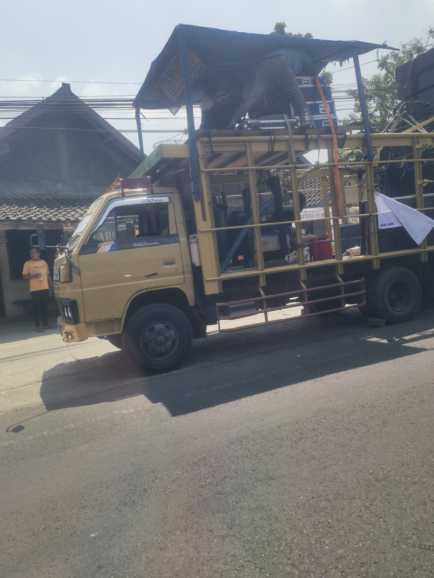A yellow Mitsubishi Colt Diesel truck is parked on a street in Tambaksari, Indonesia. The truck's bed is outfitted with a makeshift stage covered by a blue tarp, and several large speakers are visible. Two individuals are on the stage, one appears to be setting up or adjusting equipment. A man in an orange shirt stands outside a building to the left of the truck, looking towards it. The background shows residential buildings with tiled roofs and overhead power lines. The sun is shining, casting shadows on the asphalt road.