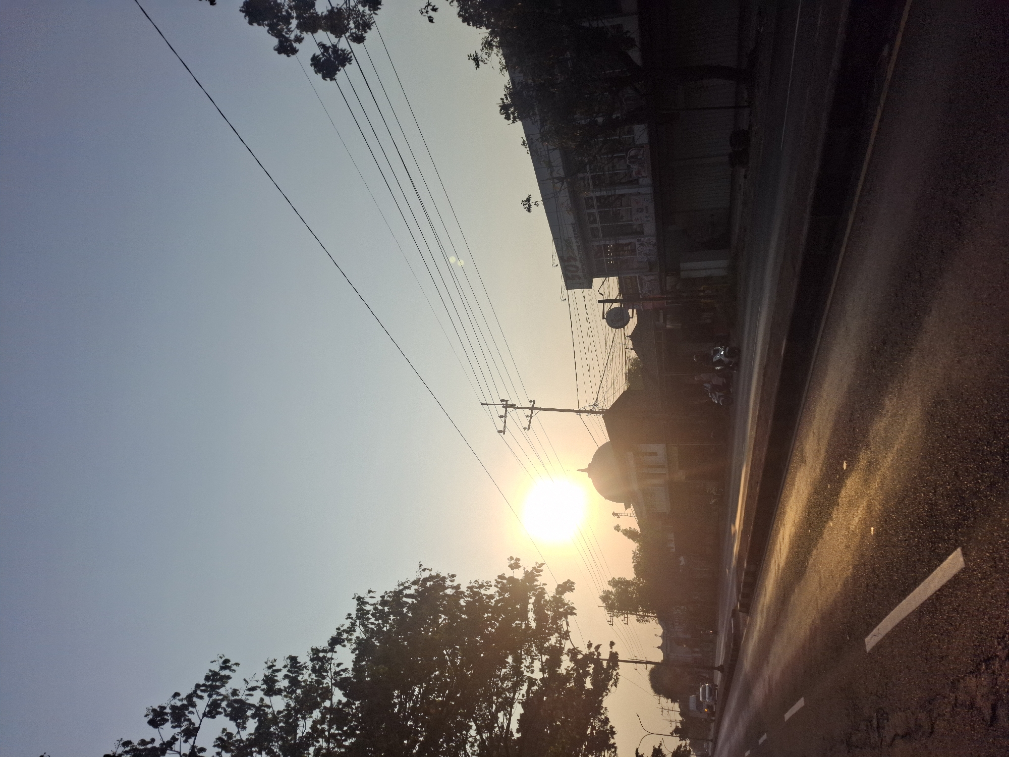 This photograph captures a street scene in Waruwetan, Indonesia, during either sunrise or sunset. The sun, a bright, luminous orb, is low in the sky, casting a warm, golden glow across the wet asphalt of the road. Reflections of the sunlight shimmer on the road's surface. Trees with dense foliage line the left side of the road, silhouetted against the sky. On the right side of the street, buildings are visible, including one with a dome, suggesting a religious structure like a mosque. Another building displays signage that reads "509 Auto," indicating a business. Utility poles with numerous wires crisscross the sky, creating a linear pattern. A few vehicles, including cars and motorcycles, are present on the road, with one motorcycle occupied by a person. The overall impression is one of a tranquil, urban setting bathed in the soft light of dawn or dusk.
