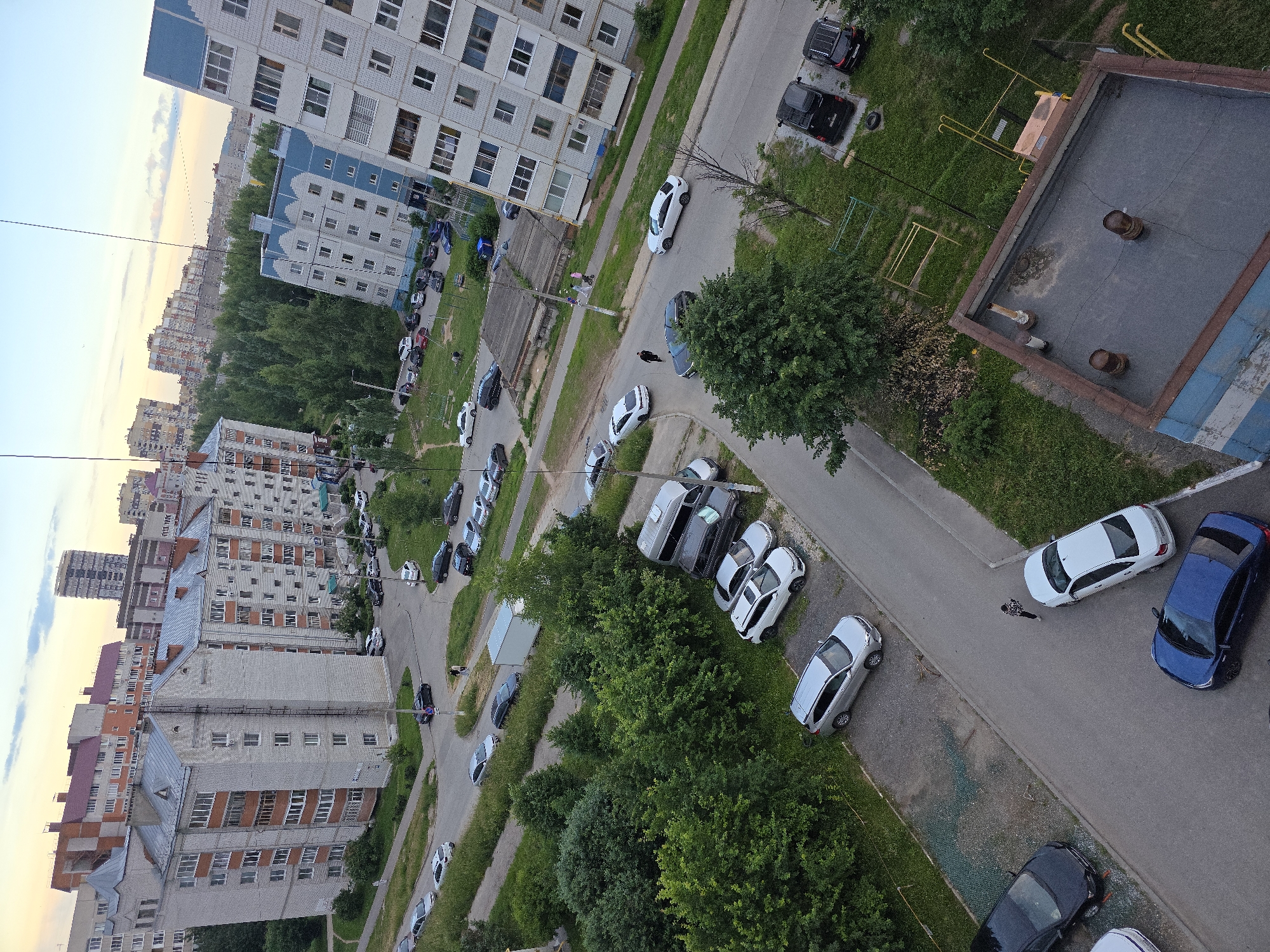 The image captures an aerial view of a residential area in Novocheboksarsk, Russia, likely during the late afternoon or early evening, indicated by the soft, warm light and the sky's coloration. The scene is dominated by multi-story apartment buildings, characteristic of Soviet-era urban planning, interspersed with green spaces and a network of roads and parking areas.
In the foreground, a flat-roofed building, possibly an industrial or commercial structure, occupies the lower right corner, showing signs of wear and tear with peeling paint and exposed pipes. Adjacent to this, several cars are parked on the street and in a parking lot, including a black SUV, a black sedan, and a dark blue sedan.
Further into the scene, a paved road winds through the neighborhood, with more parked vehicles visible along its edges. These include a silver SUV, a white sedan, and a dark colored van. A few individuals are present on the sidewalks and near the parked cars, suggesting daily life activities. Children's playground equipment, including a metal swing set, is visible in a grassy area.
The middle ground is characterized by dense green trees and shrubs, providing a natural buffer between the residential buildings. Beyond the trees, more apartment blocks of varying architectural styles and colors are visible, extending into the distance. In the far background, a hazy cityscape with taller buildings suggests the broader urban context of Novocheboksarsk. The sky above displays a gradient of pale yellow and blue with scattered clouds. The overall impression is of a typical urban residential environment in a Russian city.