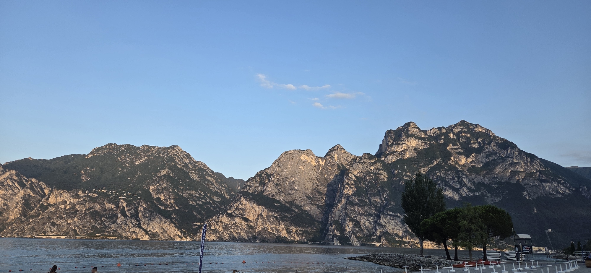 The image depicts a panoramic view of a serene lake with majestic mountains in the background, characteristic of Torbole, Italy. The water is calm and reflects the soft hues of the sky, a clear blue with a few wisps of clouds. The mountains are rugged and imposing, their rocky slopes textured with shadows and patches of green vegetation. The sunlight gently illuminates parts of the mountains, highlighting their dramatic contours.
In the foreground, the shore of the lake is visible, with a paved area and some trees planted along the edge. There are white lounge chairs arranged neatly, suggesting a recreational area. A few people are present near the water's edge, some appearing to be wading or preparing to enter the lake. A blue and white flag on a pole stands on the shore, and some orange buoys are visible in the water, marking a swimming area or a boundary.
To the right of the image, a few trees provide a touch of greenery, contrasting with the rocky landscape of the mountains. A structure that appears to be a beach umbrella stand or a kiosk is also visible. The overall atmosphere is peaceful and idyllic, capturing the natural beauty of the Italian landscape.