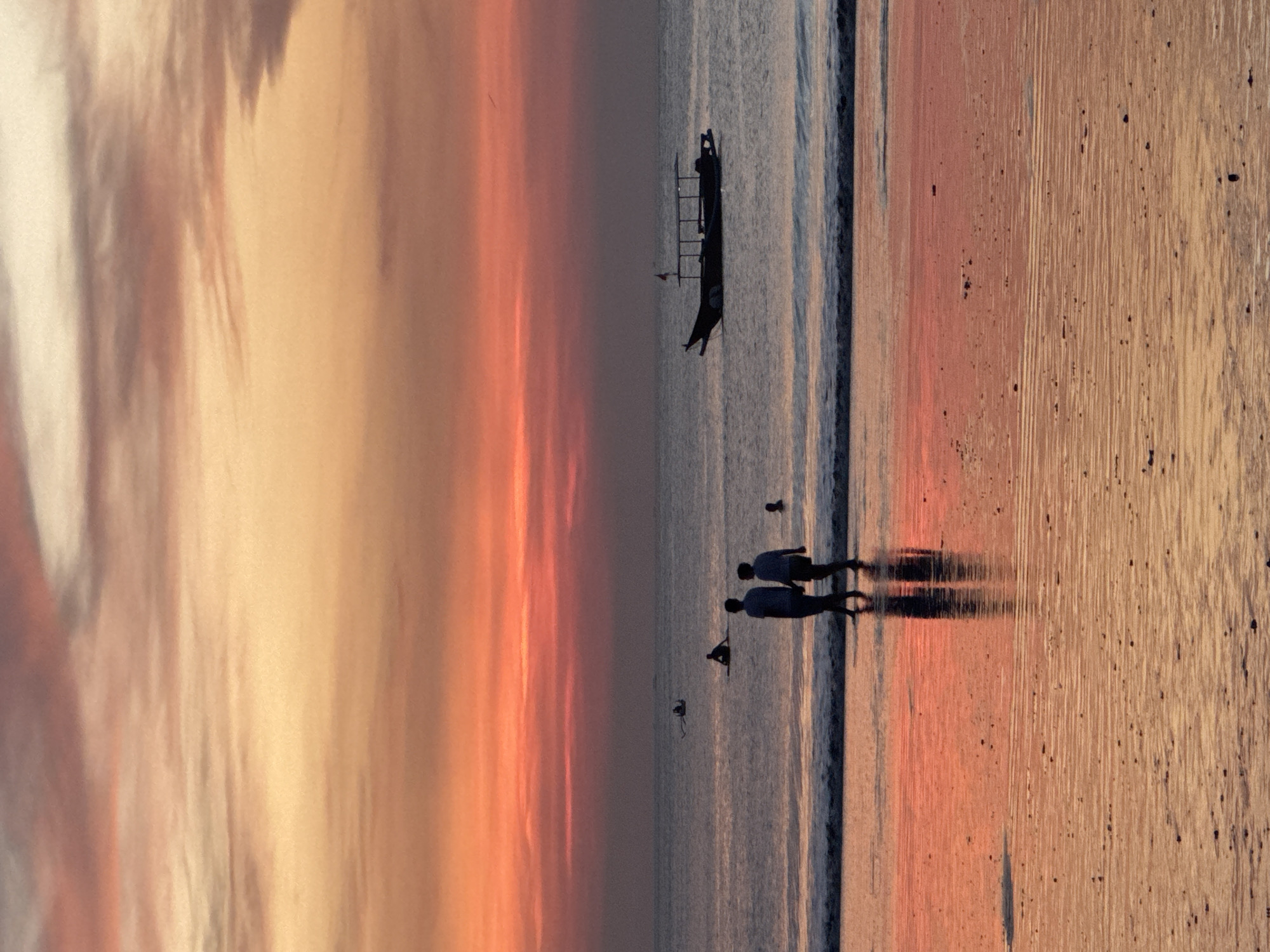 The image depicts a coastal scene, likely a beach, in Kuta Selatan, Indonesia. The sky is awash in warm tones of orange, peach, and yellow, suggesting either sunrise or sunset. The ocean reflects these colors. A traditional Indonesian outrigger boat is visible in the water. The silhouettes of two people are walking along the shoreline, with their reflections visible in the wet sand. A third person is paddling the boat. The sand has a wet, reflective surface.