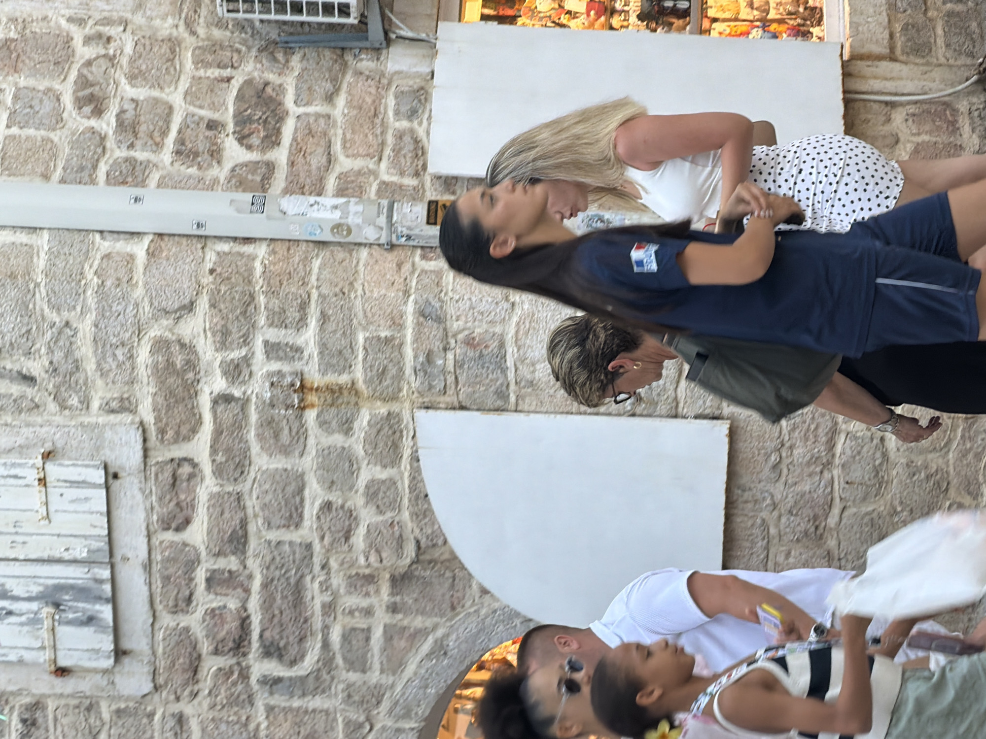 This image, taken in Budva, Montenegro, depicts a group of people in an outdoor setting with a stone-built wall as a backdrop. The perspective is from above, looking down at the subjects.
In the foreground, a young girl wearing a striped top is looking downwards, holding a white bag. Beside her, a person in a white t-shirt is partially visible.
Further up and to the left, a woman with long dark hair, wearing a dark blue t-shirt with a red, white, and blue logo, is looking to her right. Behind her, another woman with blonde hair is wearing a white top and a polka dot skirt, leaning forward.
Above them, a person with short blonde hair and glasses is visible from the shoulders up, looking to their left.
The scene appears to be during the day, possibly with warm weather, as people are dressed in casual summer attire. The stone wall suggests an old town or historical area, consistent with Budva's architecture. The overall mood seems relaxed, possibly a casual outing or shopping trip.