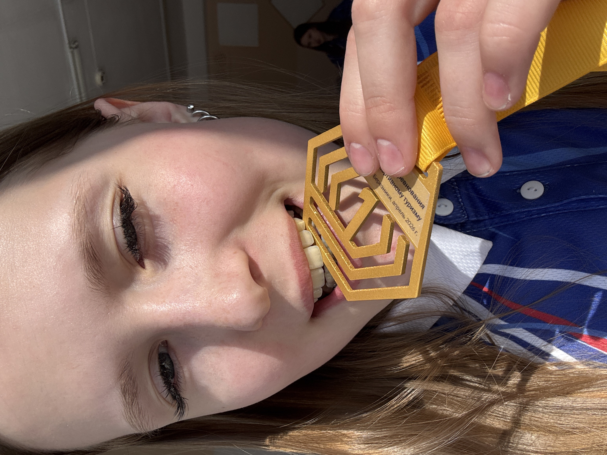 A young woman is holding a gold-colored medal in her mouth. The medal has a geometric design. The medal is suspended from a yellow ribbon. The woman has fair skin and long, light brown hair. She is wearing a blue and white striped shirt with buttons. A person is blurred in the background. The medal has text in Cyrillic including: "Березники, апрель 2026 г.", which translates to "Berezniki, April 2026." This indicates the location may be related to the city of Berezniki, which is near Usolye, Russia.