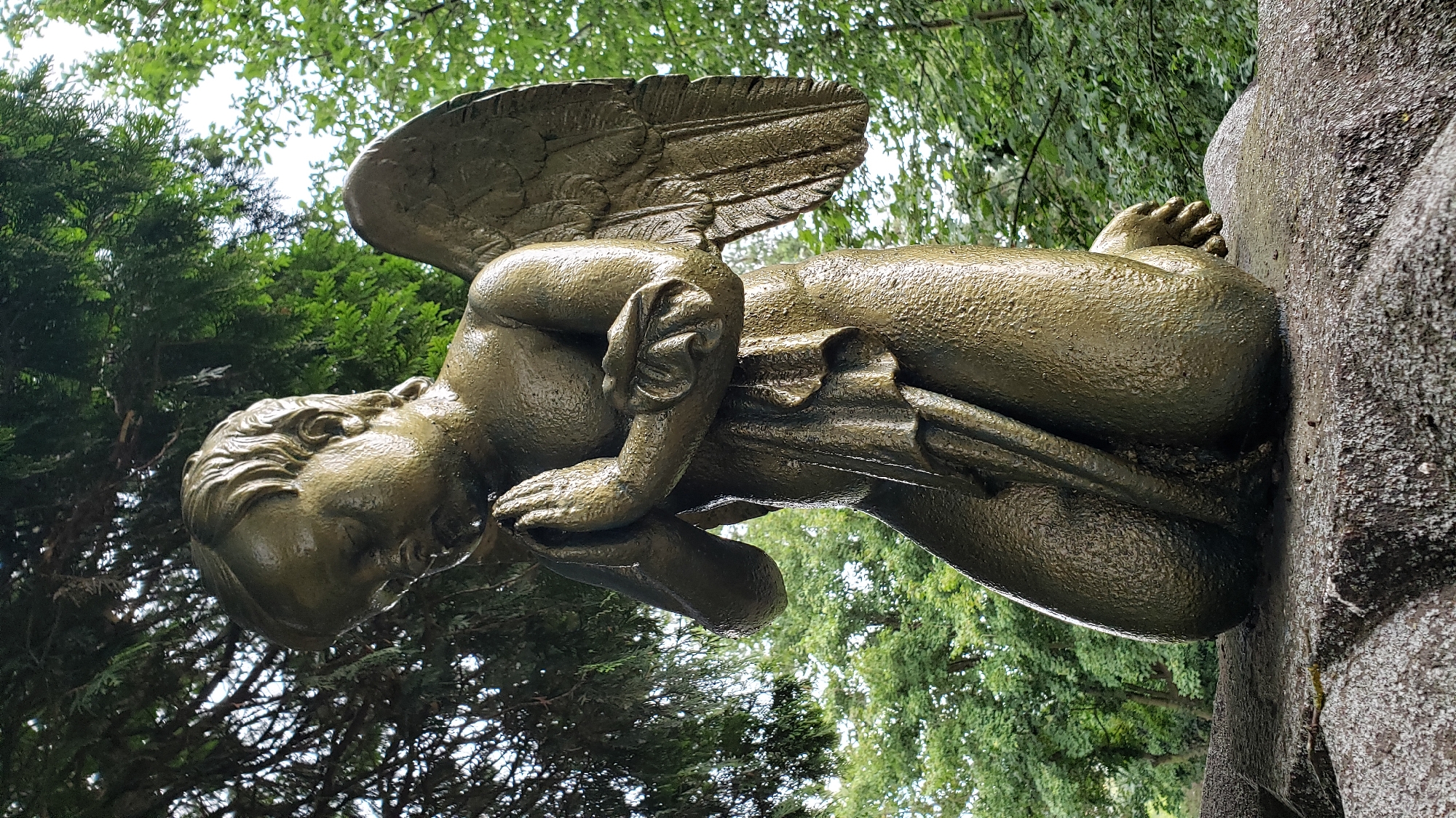 A weathered, bronze-colored statue of a young angel with wings is kneeling on a textured stone surface. The angel has its eyes closed and hands clasped in prayer. It wears a draped garment that covers its legs. The statue is situated outdoors, with lush green trees and foliage forming the background. The lighting suggests an overcast or shaded day. The location of this statue is in the Evangelical Cemetery in Prešov, Slovakia.