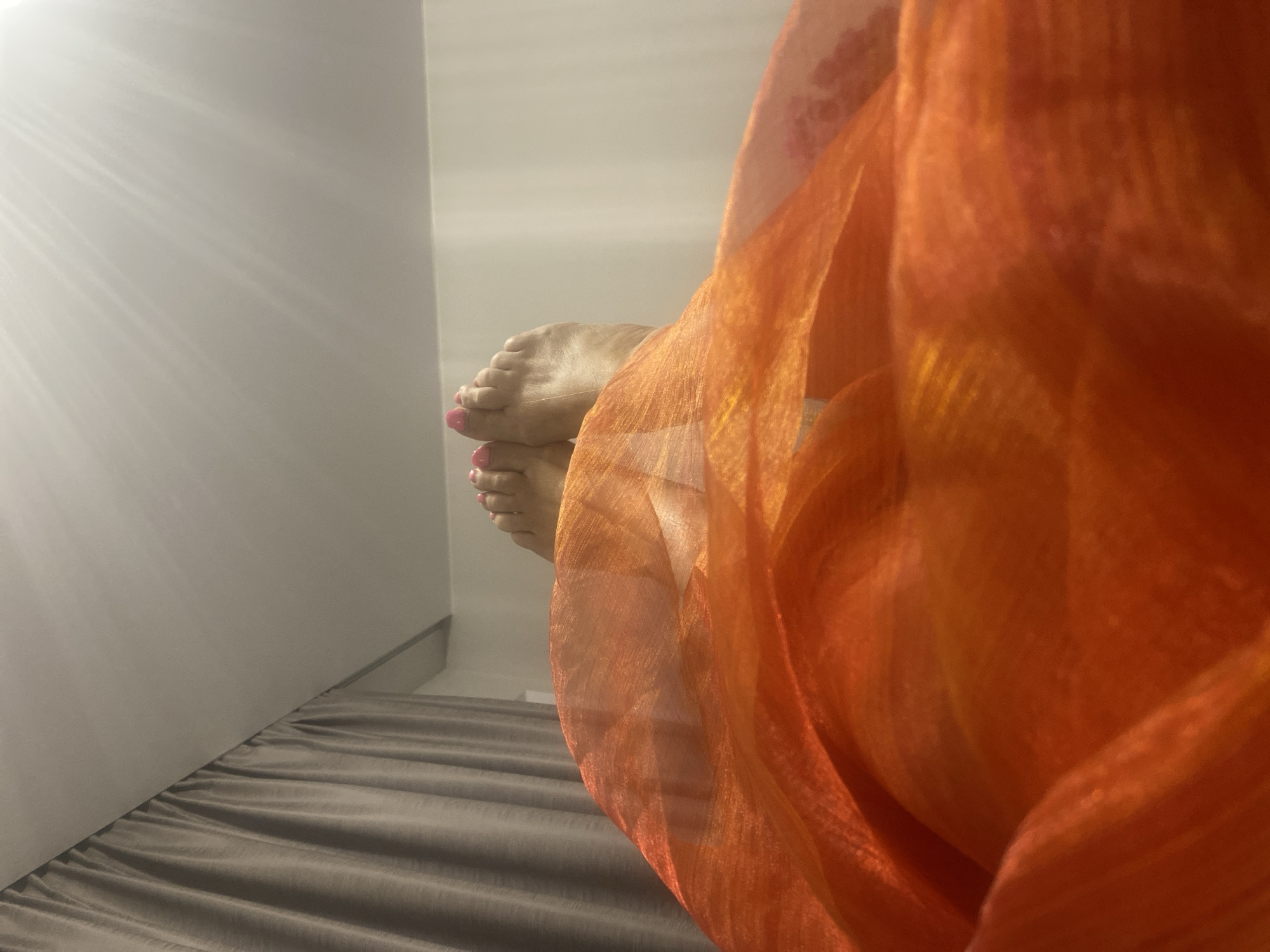 The image shows a close-up, low-angle view of a person's feet peeking out from behind an orange, sheer fabric, possibly a scarf or garment. The feet have pink nail polish on the toes. The fabric is draped in a way that creates folds and shadows, with light shining through it, highlighting its texture and color. In the background, a white wall with diagonal light streaks is visible, suggesting a light source positioned to cast these patterns. The feet are resting on a grey, textured surface, likely a bed or sofa, with visible folds in the material. The overall lighting and the soft focus on the fabric create a gentle and somewhat intimate atmosphere. No text is visible in the image. The scene appears to be indoors, possibly in a bedroom or living area. The time of day is suggested by the strong diagonal light, which could indicate sunlight coming through a window.