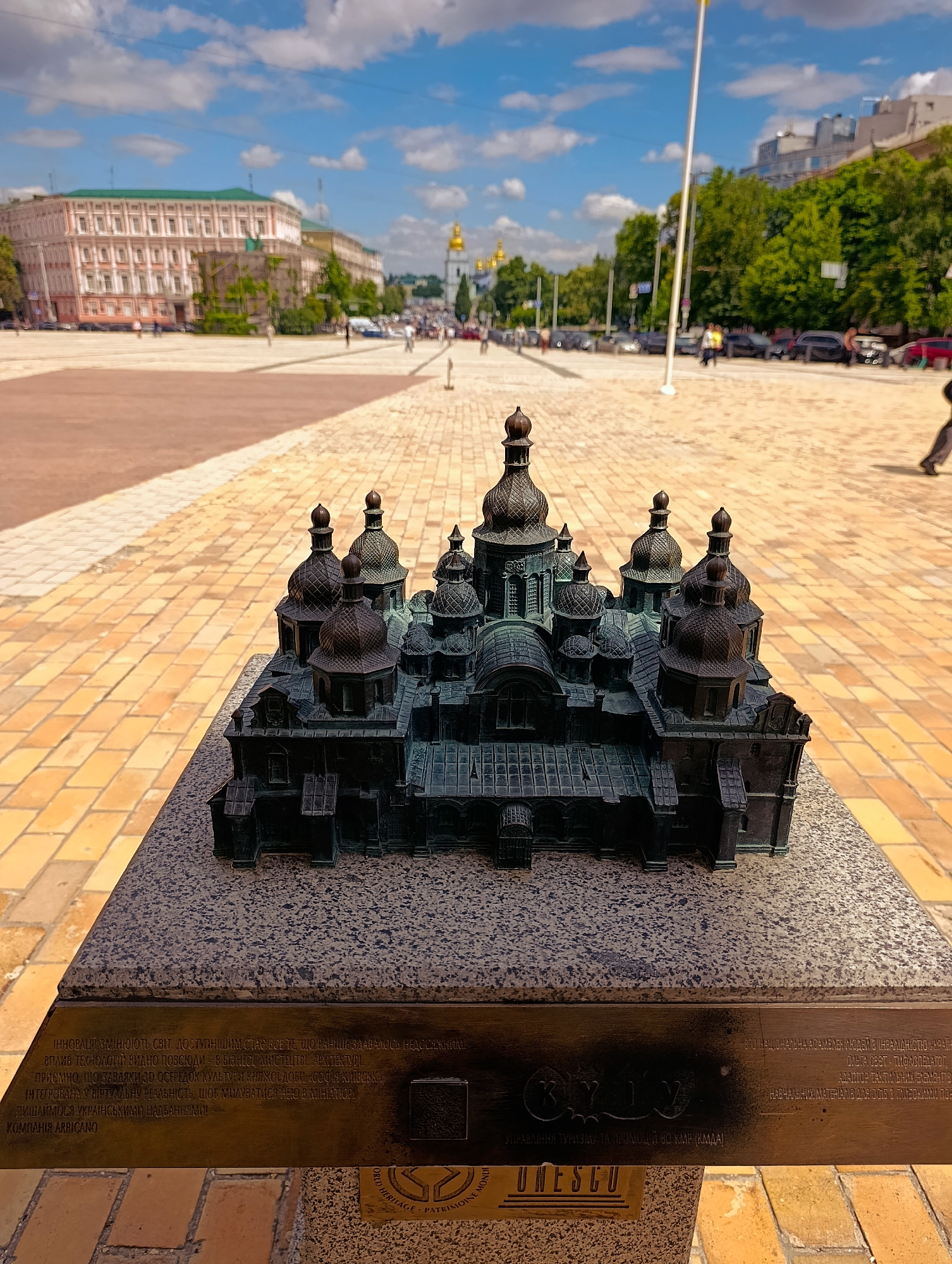 The image shows a detailed bronze miniature model of St. Sophia's Cathedral in Kyiv, Ukraine, situated on a stone pedestal. The model depicts the cathedral's distinctive domes and architectural features. In the background, a wide, paved plaza is visible, with people walking and cars on a street. Buildings, including a large pale pink building with a green roof and a golden-domed church, are also present. The sky is blue with scattered white clouds. The scene is lit by daylight, suggesting it is daytime. The inscription on the pedestal is partially visible and appears to be in Ukrainian. A UNESCO emblem is also visible on the lower part of the pedestal.