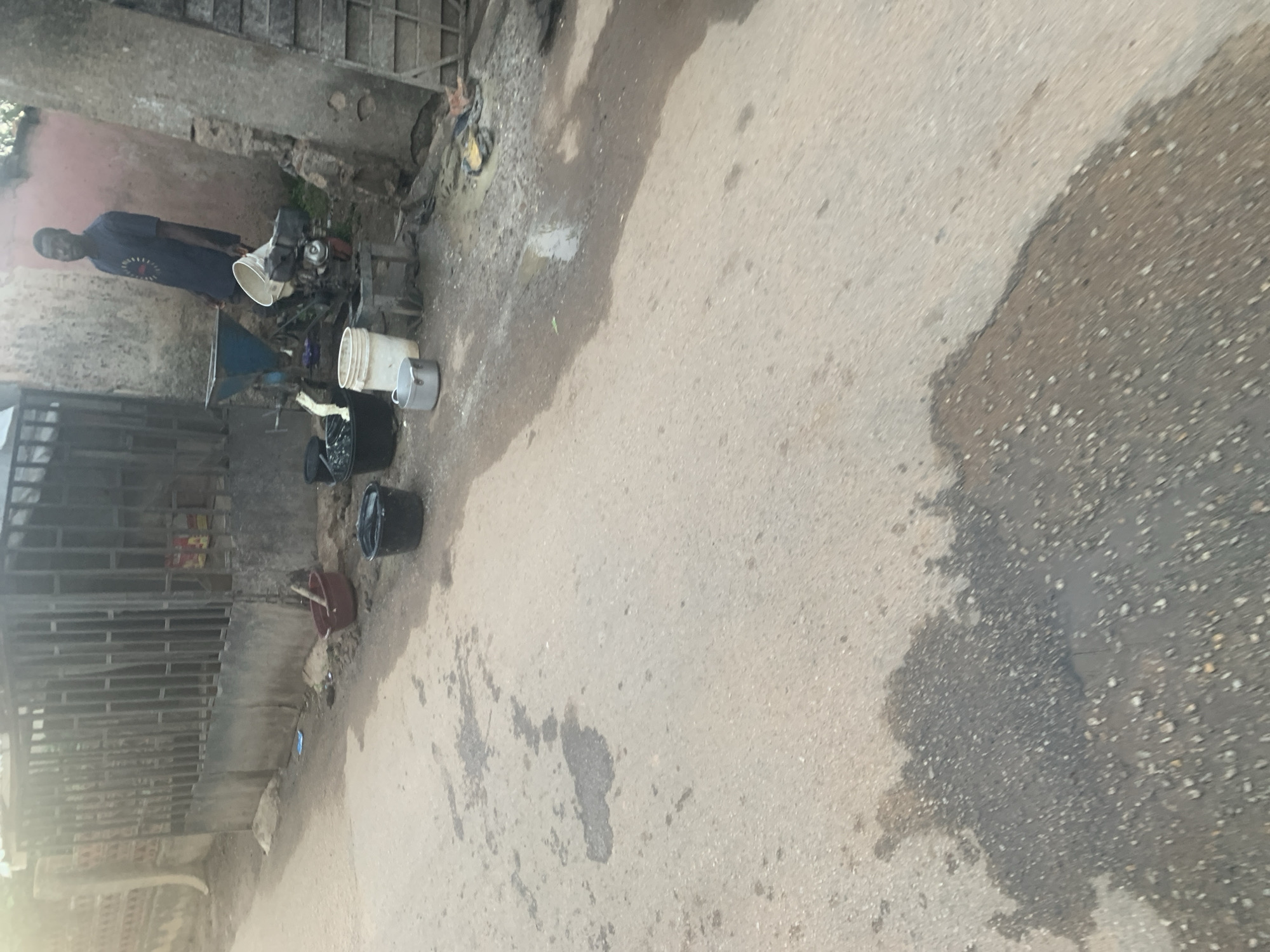The image shows a street scene in Jos, Nigeria. A man in a dark blue t-shirt stands next to a grinding machine, which appears to be powered by a small engine. Several buckets and containers are positioned around the machine, some containing a white substance that may be maize or another grain. A reddish-brown bucket with a wooden stick is also visible. The ground is paved with asphalt and shows several wet patches, possibly from recent rain or spillage. In the background, a metal fence and a rustic building are present. The overall impression is that of a small-scale processing operation taking place outdoors on a street.