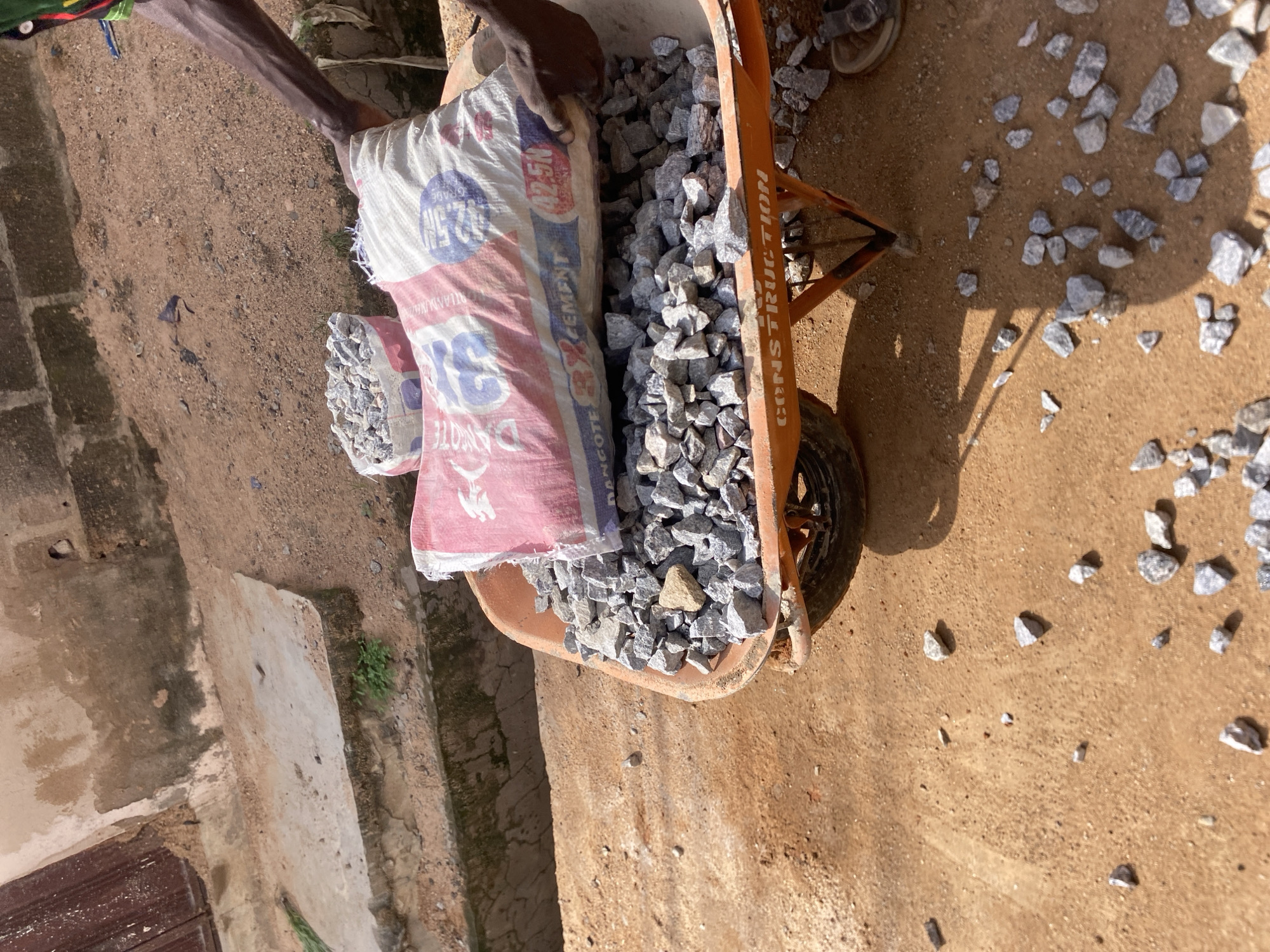 In Jalingo, Nigeria, a wheelbarrow filled with aggregate sits on sandy ground. A person's arm, visible on the right, is placing a large bag of cement, labeled "42.5N CEMENT", into the wheelbarrow. Another smaller bag of aggregate is already in the wheelbarrow. Scattered rocks and dirt surround the wheelbarrow, with a portion of a building and a trench visible in the background.