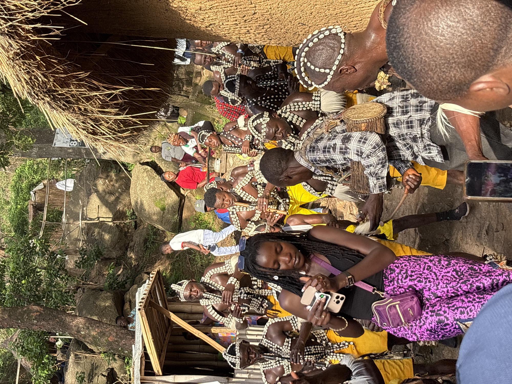 This is a photograph taken outdoors, likely in Jos, Nigeria, featuring a group of people participating in a cultural event. The scene appears to be set in a natural environment with large rocks and lush green vegetation, possibly a village or a traditional gathering place. A thatched-roof structure is visible in the background.
The primary subjects are individuals dressed in traditional attire. Many of the men are adorned with intricate necklaces and headpieces made of white shells. Some are playing wooden instruments, possibly flutes, while others are seated or resting. One man in the foreground is wearing a black and white checkered shirt over a yellow garment. A woman in the foreground is holding two smartphones and wearing a black top and a vibrant purple skirt with a black patterned design. A small purple handbag is slung across her shoulder.
The activity suggests a cultural performance or ceremony. The individuals in traditional dress appear engaged in the event, some with focused expressions, while others seem relaxed. The time of day appears to be daytime, with natural lighting illuminating the scene. The weather seems pleasant, conducive to an outdoor gathering.
Notable details include the distinct shell adornments worn by many of the men, the use of traditional instruments, and the modern presence of smartphones held by the woman, suggesting a blend of tradition and contemporary life. The overall atmosphere seems peaceful and communal. No visible text is present in the image.