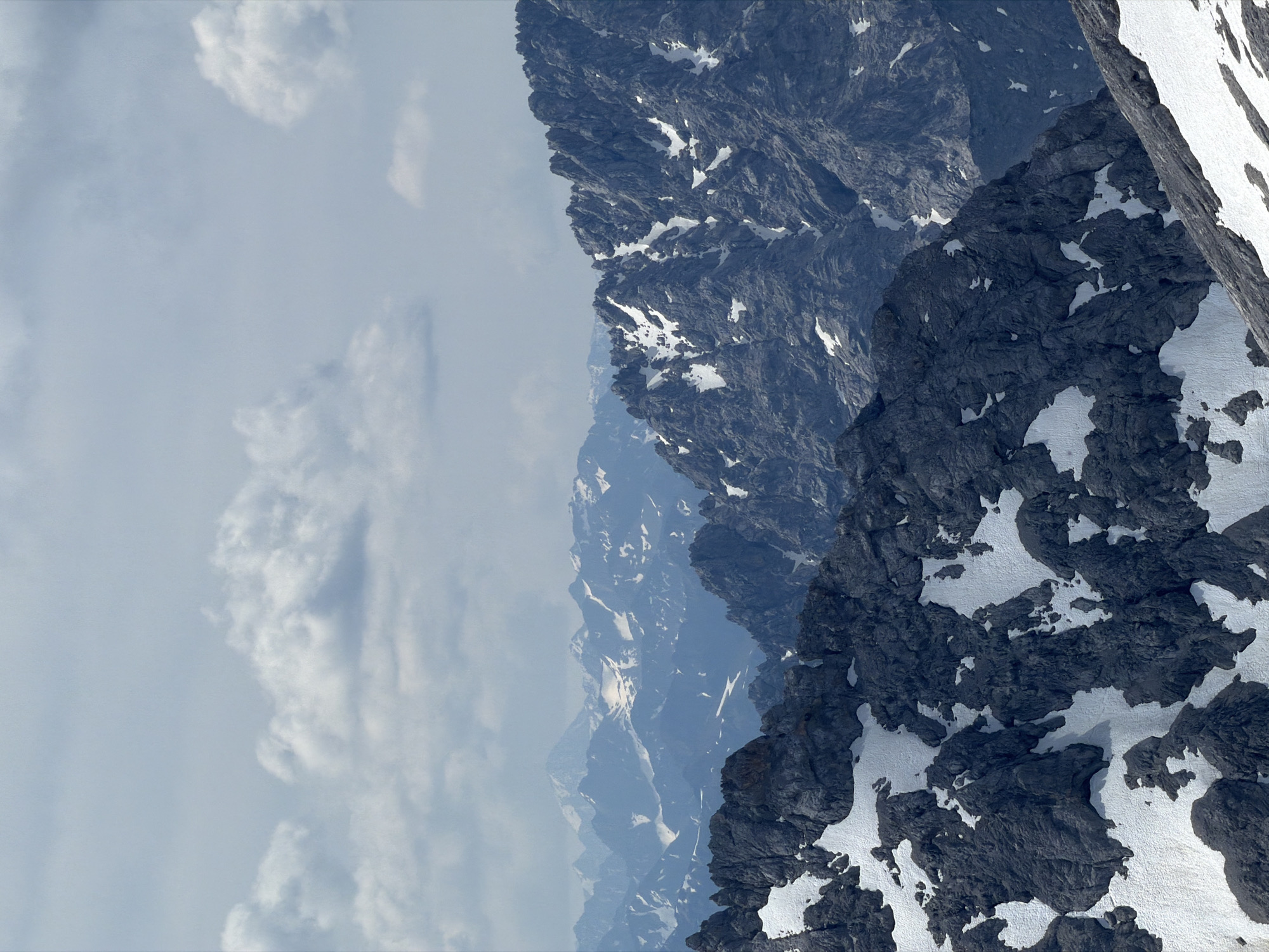 The image depicts a panoramic view of a rugged, mountainous landscape under a cloudy sky. The foreground is dominated by dark, rocky formations interspersed with patches of snow. Further back, a series of mountain ranges stretch into the distance, with varying degrees of snow cover and atmospheric haze. The scene is devoid of any human presence or man-made structures. This image likely captures a view from the Zugspitze, Germany's highest peak, located near the town of Garmisch-Partenkirchen.
