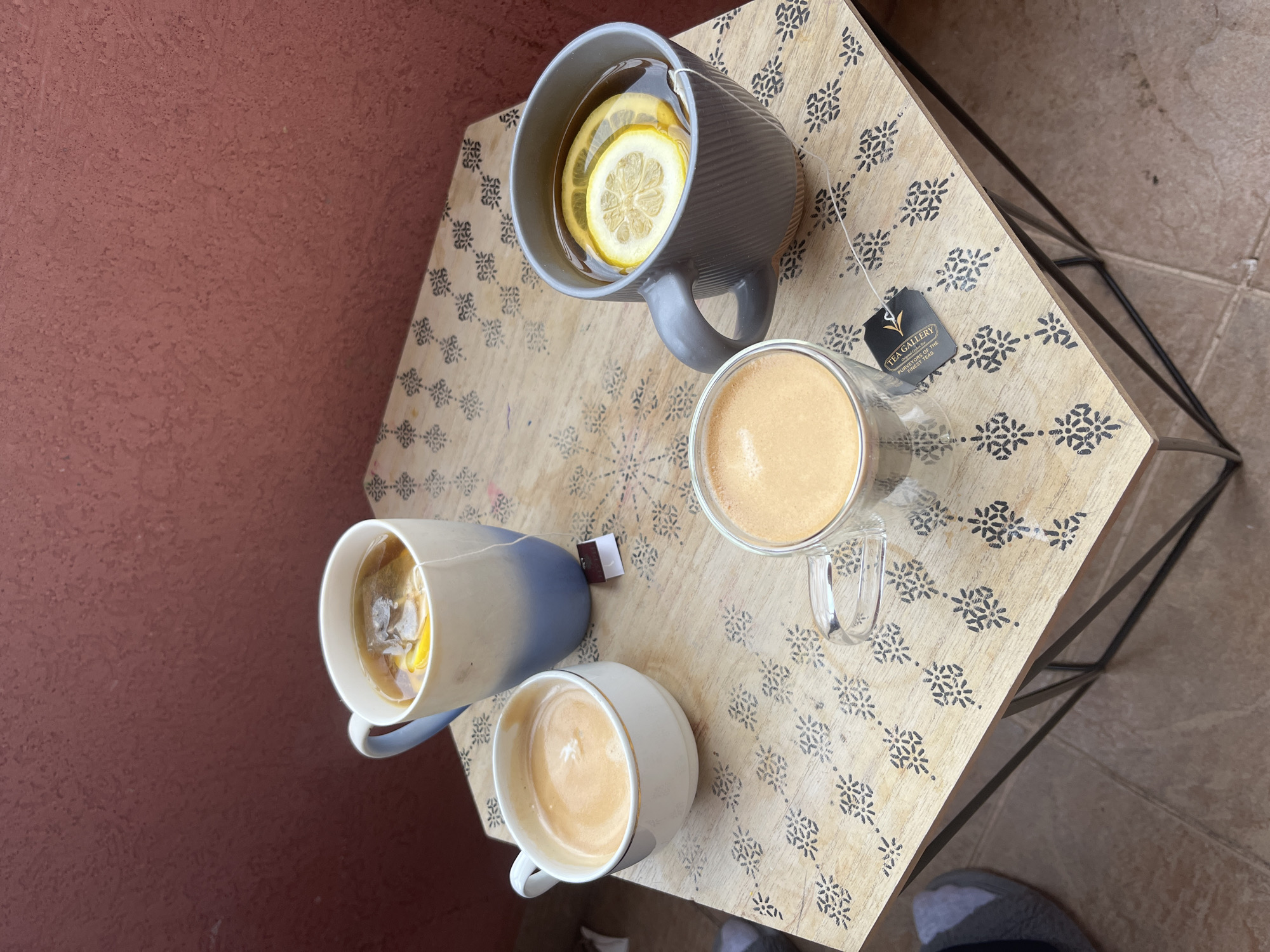 A top-down shot depicts a small, square side table with a light wood surface featuring a dark floral pattern. Four beverages are arranged on the table: a gray mug filled with hot tea and lemon slices, a blue and white mug containing tea and lemon, a clear glass mug with coffee, and a white mug with coffee. A tea bag tag is visible near the clear mug. The table sits against a dull, pink wall on one side and a tiled floor on the other. The setting is likely a cafe or patio, with a focus on the assortment of drinks. The location in the city of Budva, Montenegro is unspecified and cannot be determined from the image.