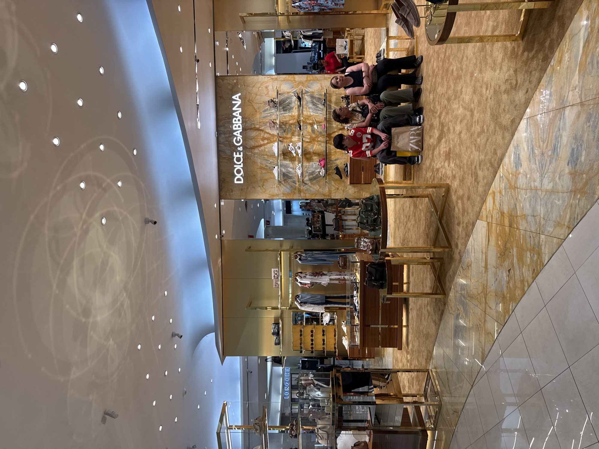 The image depicts the interior of a Dolce & Gabbana store, likely within a high-end shopping center. The store's facade features the brand name in capital letters. Inside, shelves display shoes and other merchandise. Several people are seated on a bench near the entrance. A boy wearing a red and white sports jersey is sitting next to a shopping bag from Zara. There is no evidence in the image to suggest the location is in Puebla, Mexico. The interior design includes marble flooring, gold-colored metal display stands, and a bright, white ceiling with recessed lighting. The overall impression is of a luxurious and well-lit retail environment.