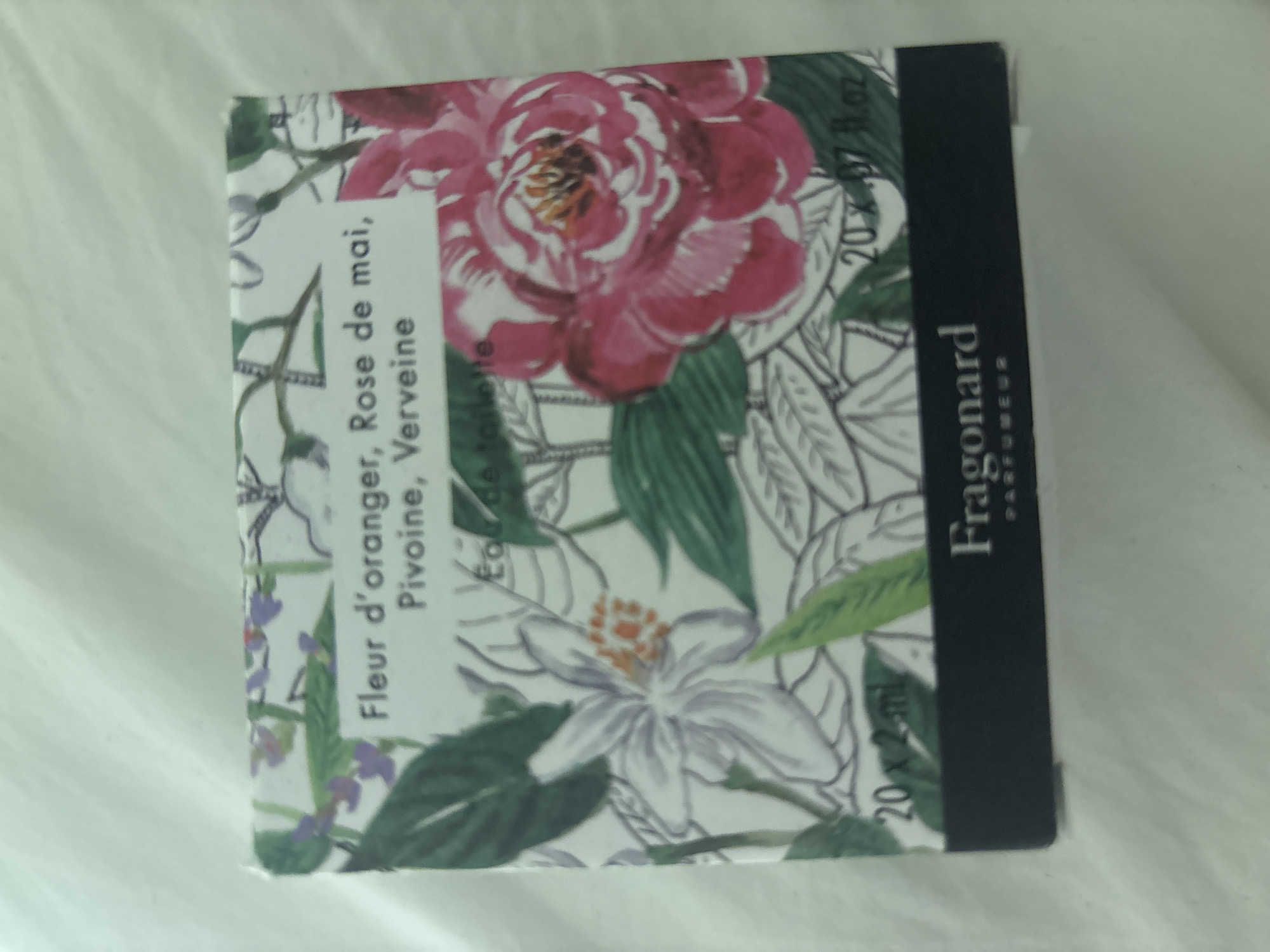 The image shows a close-up of a box of Fragonard perfume. The box features a floral design with pink roses and white orange blossoms, as well as green leaves. The text on the box indicates the fragrance notes are "Fleur d'oranger, Rose de mai, Pivoine, Verveine" and it is an "Eau de Toilette". It also states "20 x 2ml" and "20 x .07 fl.oz", suggesting it is a multi-pack of small perfume vials. The brand name "Fragonard" and "PARFUMEUR" are visible at the bottom of the box. The box is placed on a white, textured surface, possibly fabric. There is no discernible connection to Dubai, United Arab Emirates in the image.