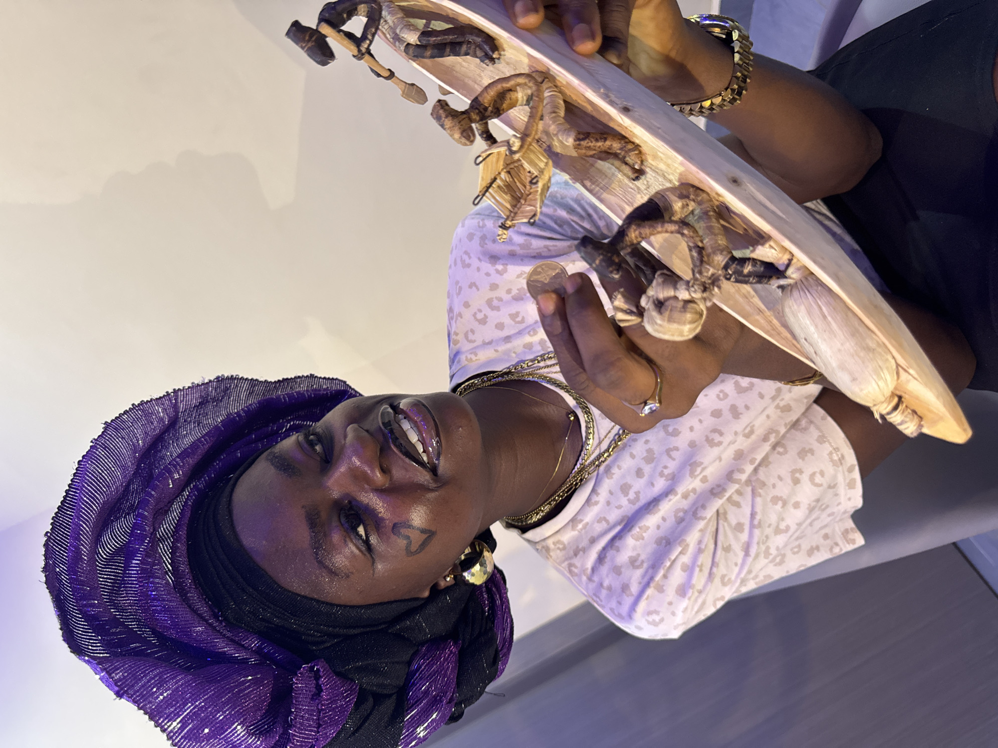 A woman in Ajara, Nigeria, holds a wooden platter with small, stylized wooden figures of people performing various tasks. She is wearing a purple head wrap, a white patterned shirt, and gold jewelry. She has a heart drawn on her left cheek. She holds a coin in her left hand.