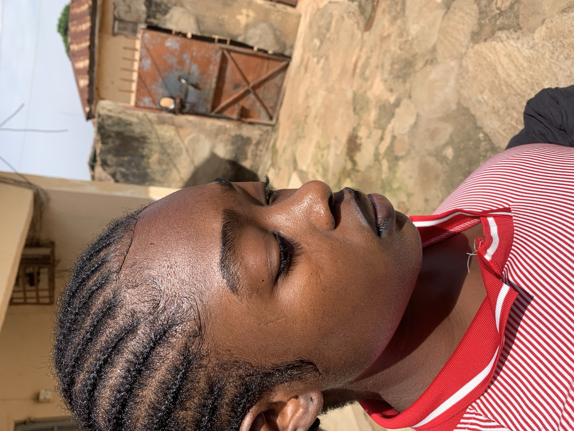 The media file shows a close-up, side-profile view of a young Black person with their eyes closed, looking upwards. They have dark skin, dark braided hair styled with cornrows, and are wearing a red and white striped shirt. The lighting suggests it is daytime, with harsh shadows indicating bright sunlight.
The background is out of focus but appears to be an outdoor setting with textured, earthy-colored walls and a rustic, weathered metal door with crisscross bracing. The ground is also textured, possibly a rough stone or concrete surface. A bit of greenery can be seen peeking from the top left of the frame.
The person's facial expression is serene, suggesting rest, meditation, or sleep. The overall scene conveys a sense of peace and stillness, possibly in a warm, sunny environment. The location context provided is Rafin Par, Nigeria. No visible text is present in the image.