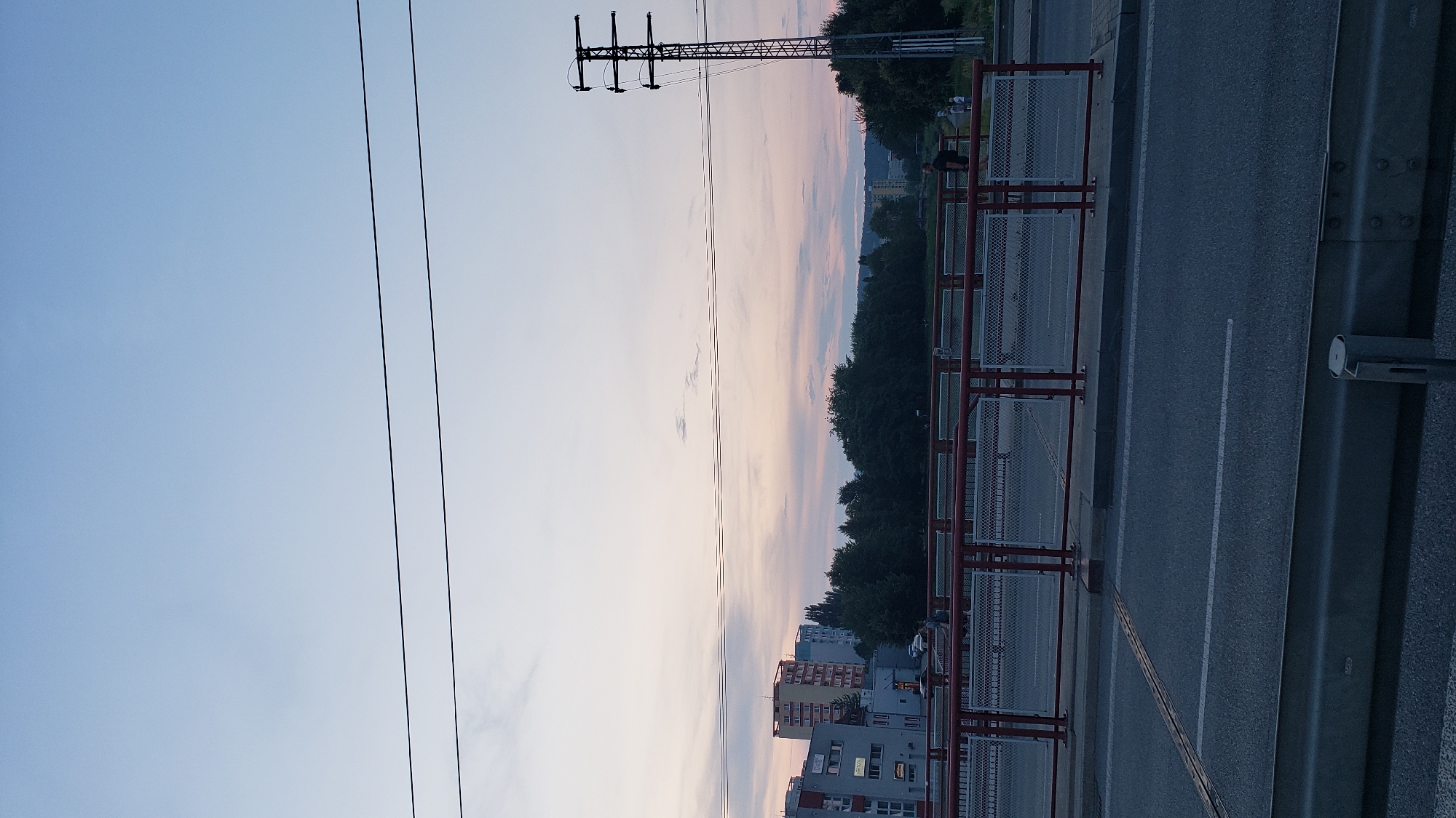 The image depicts a scene in the city of Prešov, Slovakia, captured during twilight. The sky is a gradient of light blue to pale pink, indicating the end of the day, with a few wispy clouds scattered across it. Several parallel power lines stretch horizontally across the upper portion of the frame. In the middle ground, a red-railed pedestrian bridge with metal mesh panels spans across. Beyond the bridge, a line of trees forms a dark green barrier, partially obscuring buildings in the distance. On the right side of the image, a tall, industrial-looking electricity pylon stands out against the sky. The foreground shows a paved road with white lane markings, suggesting it is a street or a bridge deck. Several indistinct figures, presumably pedestrians, can be observed on the bridge. The overall atmosphere is calm and subdued, characteristic of an urban setting at dusk.