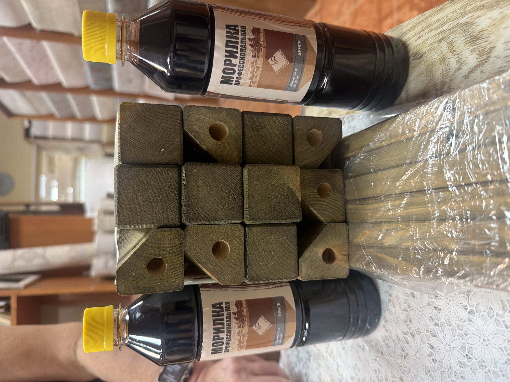 In this image, two bottles of "MORILKA PROFESSIONAL" wood stain, labeled in Russian as "МОРИЛКА ПРОФЕССИОНАЛЬНАЯ" and indicating a volume of 0.5L, are placed on top of a stack of wooden posts. The bottles have yellow caps and dark liquid. The wooden posts are square-shaped, with holes drilled through some of them, and are wrapped in clear plastic. The label on the bottles shows a landscape illustration and the brand name "LIGNUM ARMOUR". The text "ВЕНГЕ" likely refers to the color or type of stain. The scene is indoors, suggested by the shelving and indoor lighting visible in the blurred background, and the presence of a lace-covered surface beneath the objects. The overall impression is one of products prepared for use or storage.