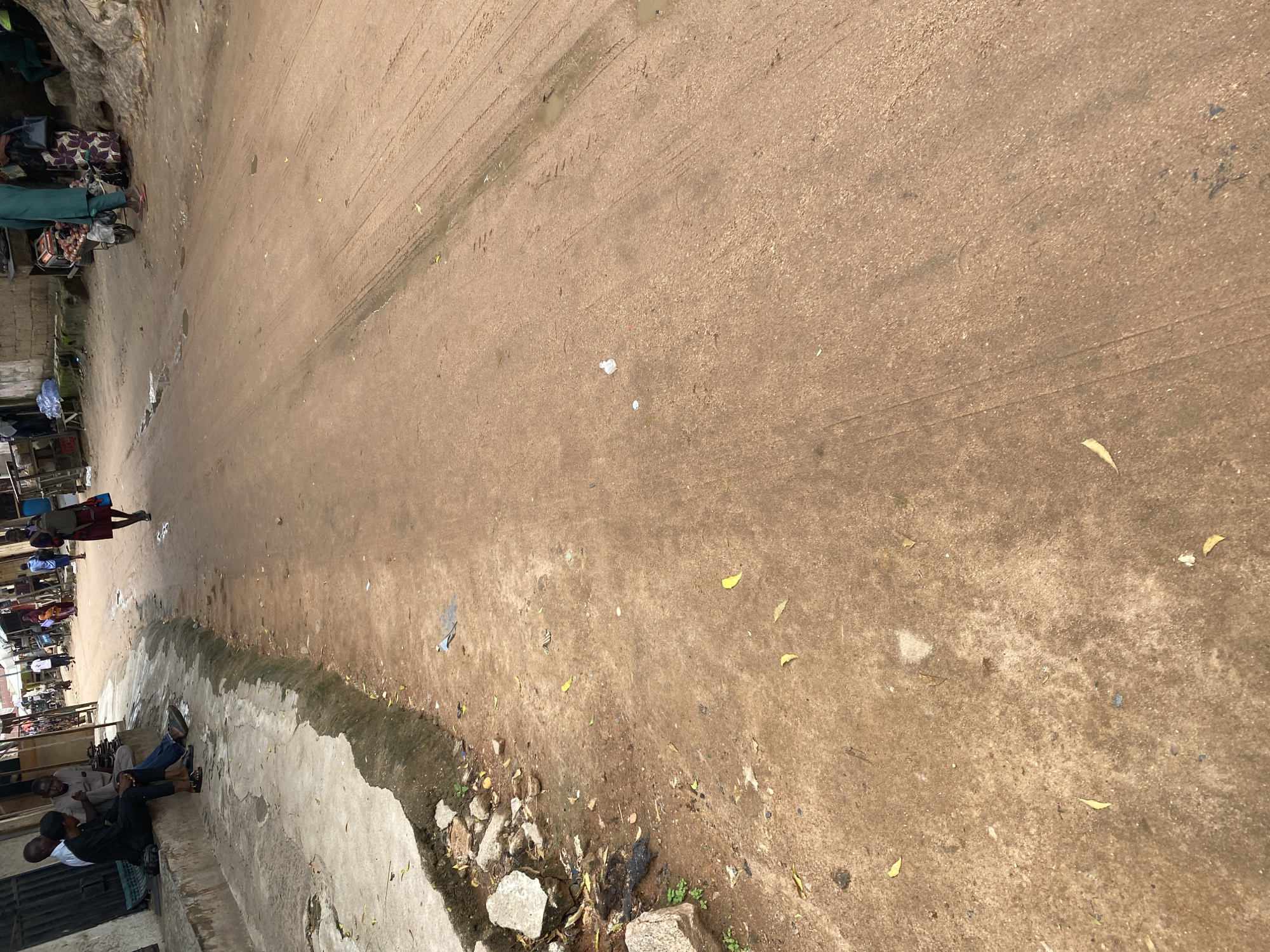 The image depicts a street scene in Jalingo, Nigeria. The ground is unpaved, dusty, and shows tire tracks from vehicles, with scattered debris and puddles of water. A concrete sidewalk runs along the left side of the road, where several people are seated or standing.
On the sidewalk, a group of men are gathered, some appearing to be in conversation. One man in a black shirt and dark pants is seated, looking towards another man. Further down the sidewalk, more individuals are visible, some near market stalls or small shops. A person in a dark red top is walking away from the camera on the road.
The scene suggests a busy, everyday market or street environment. The lighting indicates it is daytime, possibly late morning or early afternoon, given the shadows. The weather appears to be humid or recently rained, evidenced by the puddles and dampness on the ground. The overall atmosphere seems casual and communal, with people engaged in daily activities and social interactions. There is no visible text in the image.