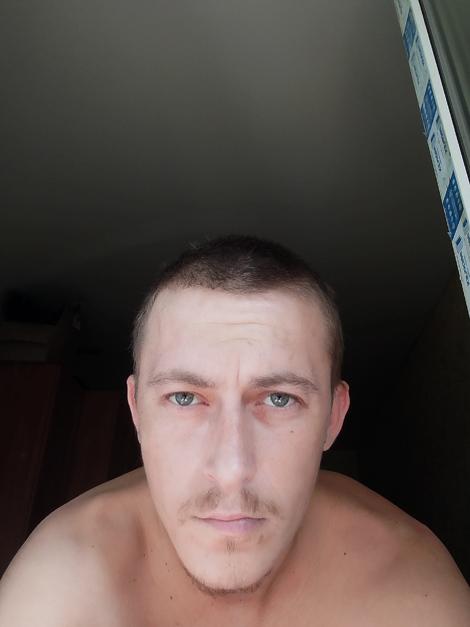 The image shows a close-up, head-and-shoulders selfie of a Caucasian male with short, dark hair and green eyes. He has a light mustache and stubble on his chin. The man is looking directly at the camera with a neutral expression. His shoulders are bare. The background is dark, with a hint of a window frame on the right side of the image. The window frame has a blue and white sticker with text on it. There is also a dark wooden furniture item visible in the lower left portion of the background. The image appears to have been taken indoors. There is no information in the image to suggest a location in the city of Ушья, Russia.