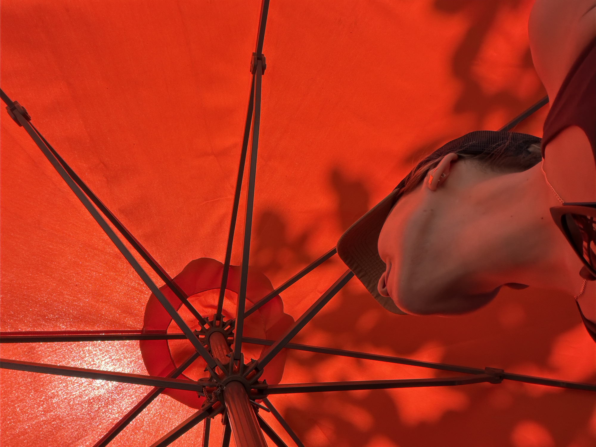 The image shows a person seen from below, inside an open orange umbrella. The umbrella's framework of dark metal ribs is visible, extending towards the canopy. The person is wearing a brimmed hat and their face is turned away from the camera, with only their ear, neck, and the side of their head visible. A strap from what appears to be a bag or clothing is visible on their shoulder. The bright orange of the umbrella fabric is the dominant color, with sunlight filtering through, casting shadows on the canopy. The scene appears to be outdoors, likely during the day, given the strong sunlight. The overall impression is one of seeking shade or shelter. No text is visible in the image.