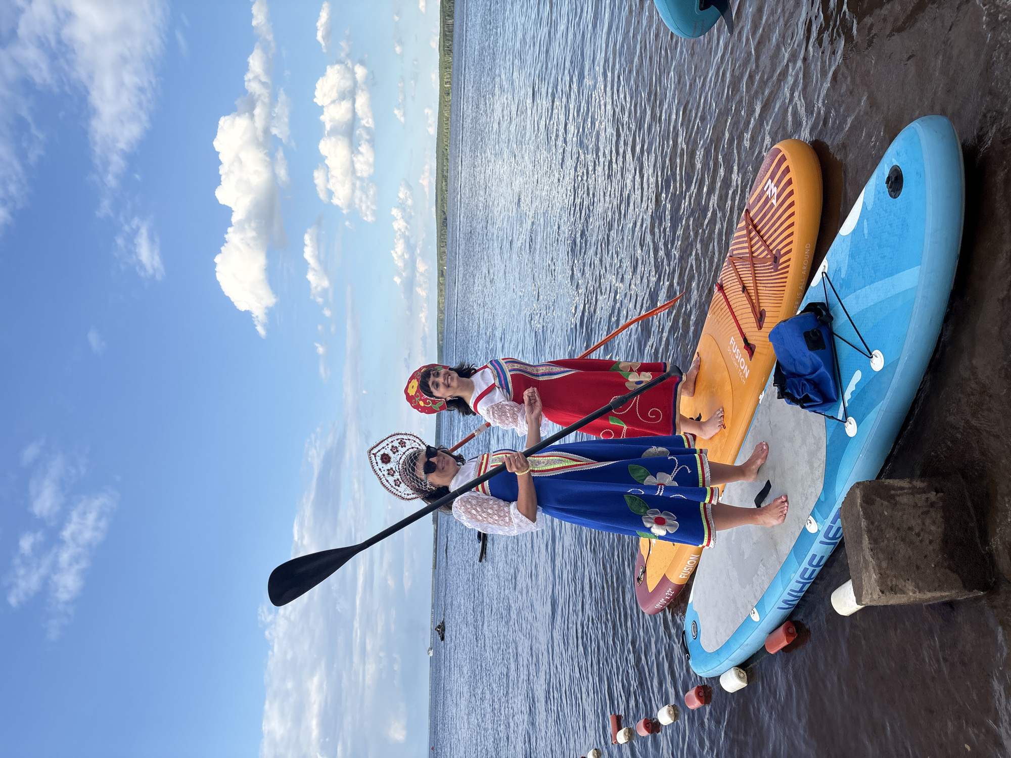 In Chaikovsky, Russia, two women dressed in traditional, vibrant costumes stand on paddleboards in the water. The woman in front wears a blue dress with embroidered white flowers and a decorative headpiece, while the woman behind her is in a red dress with similar embroidery and a red head covering. Both women are smiling and appear to be enjoying themselves. They are holding paddles, suggesting they are about to embark on or have just finished paddleboarding.
The scene is set against a backdrop of a large body of water, likely a river or lake, with a tree-lined horizon visible in the distance. The sky is bright blue with fluffy white clouds, indicating a clear and pleasant day, likely midday. The paddleboards are bright blue and orange, with some text visible on them, including "FUSION" and "WHEE JEZ". There are also buoys floating in the water near the paddleboards. The overall atmosphere is cheerful and picturesque, showcasing a cultural experience in a natural setting.
