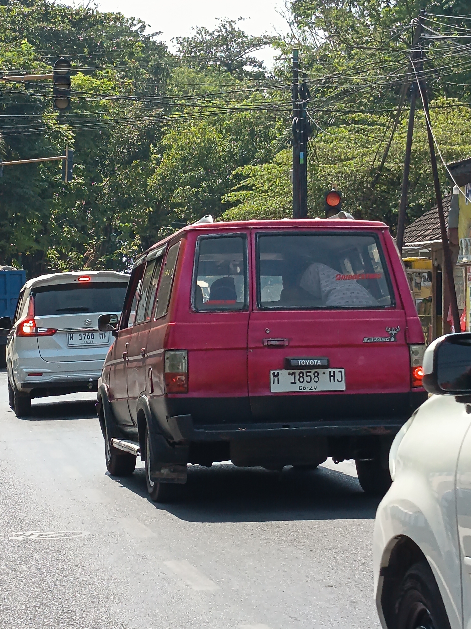 The image shows a street scene in Kandangjati Kulon, Indonesia, on a sunny day with dense foliage in the background. A red Toyota Kijang is in the foreground, facing away from the camera. The vehicle has a license plate that reads "M 1858 HJ" and the year "06-20". Inside the car, a person wearing a hijab and a patterned shirt is visible. To the left of the red car, a white Suzuki Ertiga is also present, with its license plate reading "N 1768 MF" and "06-21". Traffic lights are visible in the background, with one showing an orange light. Power lines and poles are also prominent in the environment. The road surface appears to be asphalt.