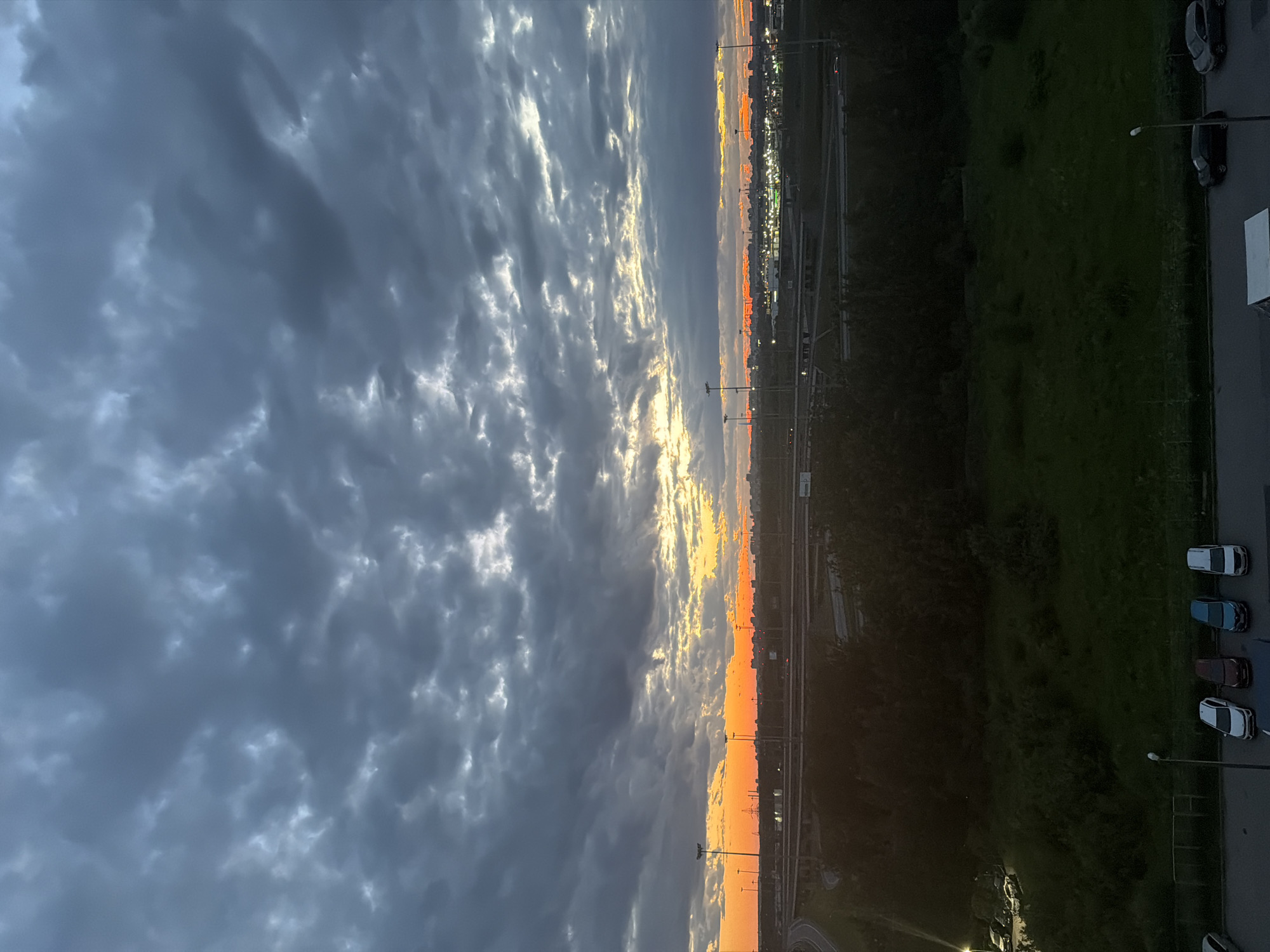 The image captures a dramatic sky at dusk over a city. The sky is filled with heavy, dark clouds, with streaks of orange and yellow light breaking through near the horizon, indicating the setting sun. Below the sky, a complex network of highway overpasses and underpasses stretches across the landscape, suggesting an urban or suburban environment. Patches of dark green foliage and trees are visible between the highway structures and in the foreground. In the lower part of the image, a parking area is shown with several cars parked. The overall scene is one of a transitioning day, with the fading light casting long shadows and creating a somewhat moody atmosphere.