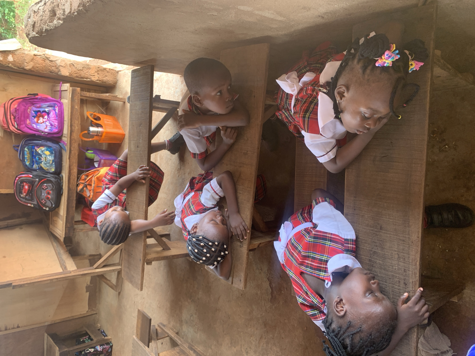 In a classroom in Ukwu-Abwa, Nigeria, five young children in red and white plaid uniforms are resting their heads on wooden desks. Backpacks hang on a shelf in the background. The classroom appears to be constructed with simple wooden supports and earthen walls.