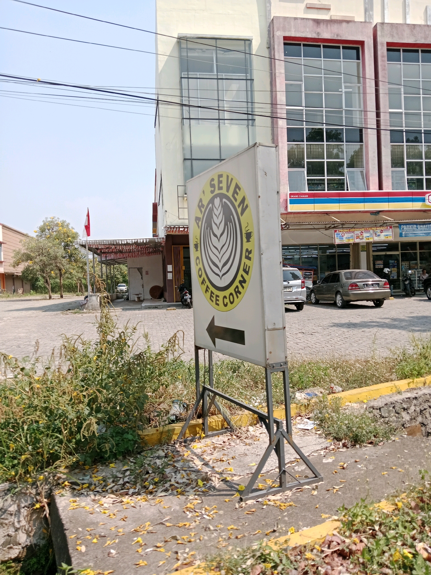 A sign for "Ar' Seven Coffee Corner" stands prominently in the foreground, angled towards the right. The sign is rectangular and white with a circular logo depicting a coffee cup and leaves. An arrow points to the right on the lower half of the sign. The sign is supported by a metal frame. The ground in front of the sign is covered with fallen yellow leaves and dry grass.
Behind the sign, a paved area is visible, with a light-colored car and a dark sedan parked. In the background, a multi-story building with large windows dominates the scene. A red and white Indonesian flag flies from a pole on a covered outdoor area to the left of the building. Trees with yellow flowers are present in the mid-ground. Power lines are strung across the sky. The environment appears to be a street or parking lot in Sabandar, Indonesia, possibly in front of a commercial establishment.
