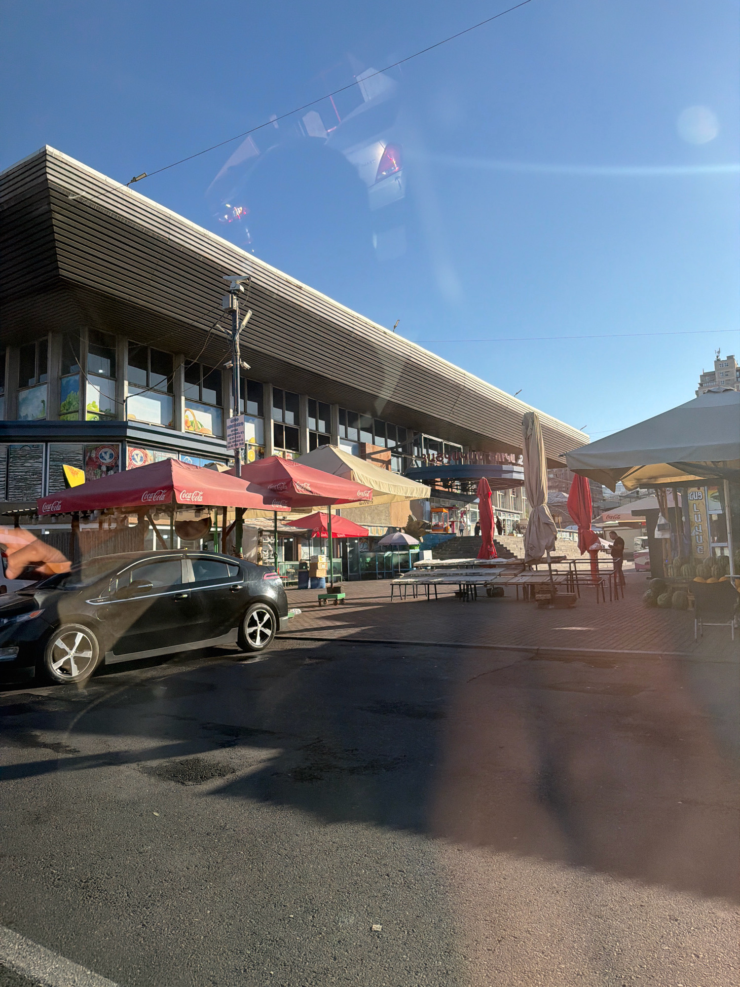 The image depicts an outdoor market scene under a clear blue sky. In the foreground, a black car is parked on asphalt, with its tires and wheels visible. The car is facing left. Behind the car, a row of red and beige market umbrellas are set up, some with "Coca-Cola" branding. Tables and chairs are arranged in front of these stalls.
The background features a large, modern building with horizontal slatted elements and glass windows. Further back, more market stalls are visible, some selling produce such as watermelons. A man is standing near one of the stalls, looking down. A few people can be seen walking in the distance. The overall impression is of a bustling marketplace on a sunny day.
The location of this market is the Vernissage market in Yerevan, Armenia, known for its arts, crafts, and souvenirs.